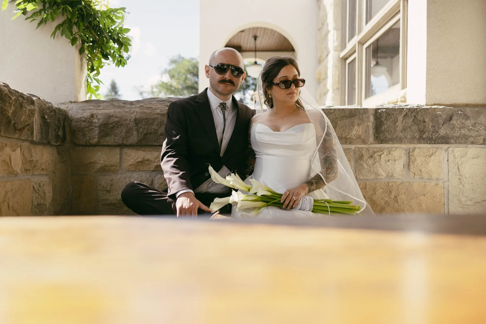  A bride and groom wearing sunglasses sit closely together on a stone bench. The bride holds white flowers and wears a white dress and veil, while the groom is in a dark suit with a tie. Sunlight fills the outdoor setting. 