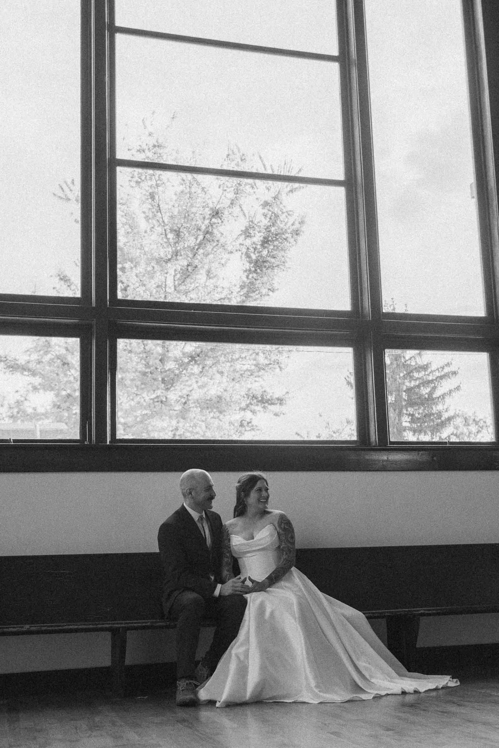  A bride in a flowing white gown and a groom in a suit sit closely together on a bench beneath a large window inside TRICA, smiling at each other. Natural light fills the room and trees are visible outside. 