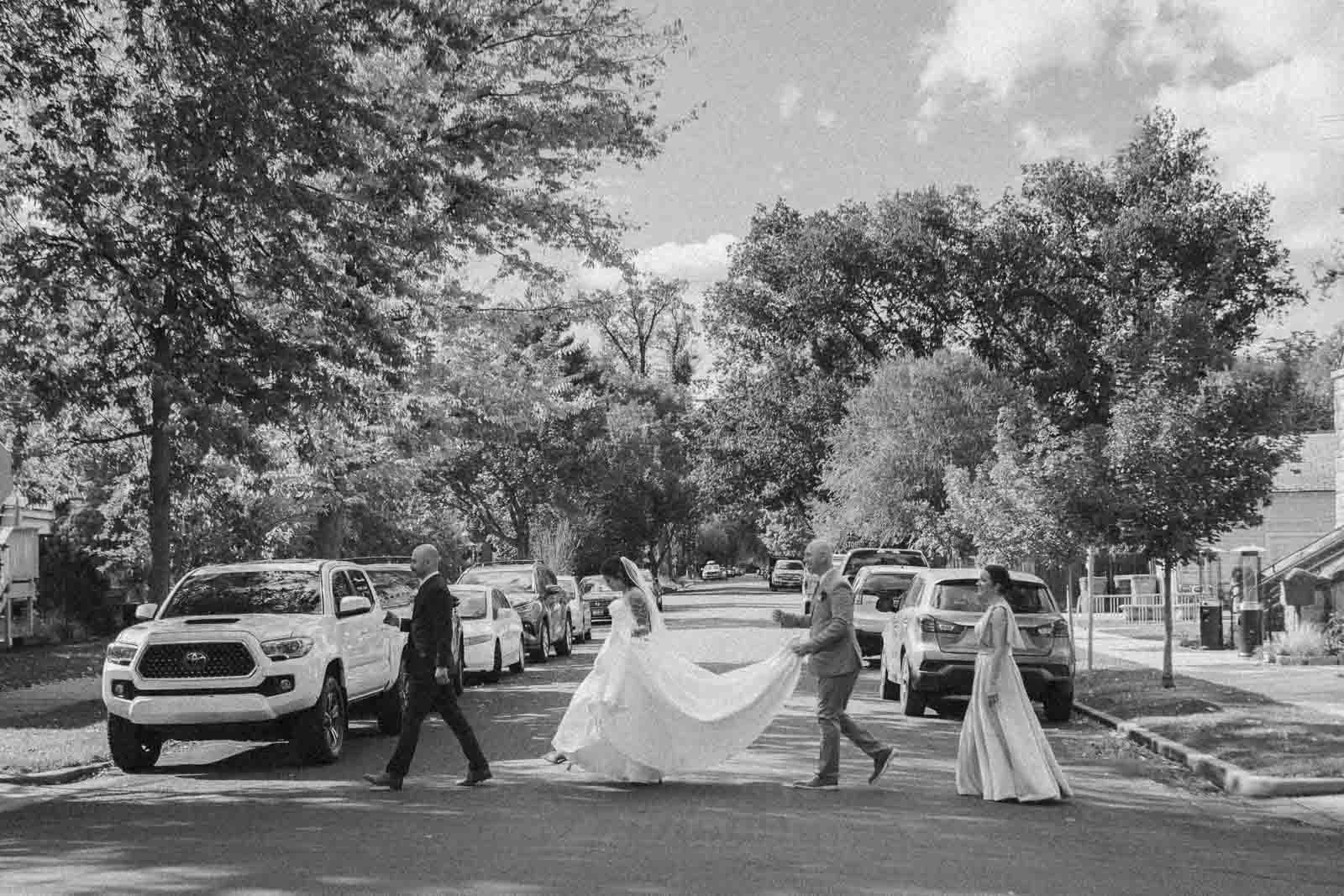  A bride in a flowing gown crosses a tree-lined street, assisted by three people, including a person holding her dress. Parked cars line both sides of the street on a sunny day. It echoes to Abbey Road studio cover by The Beatles 