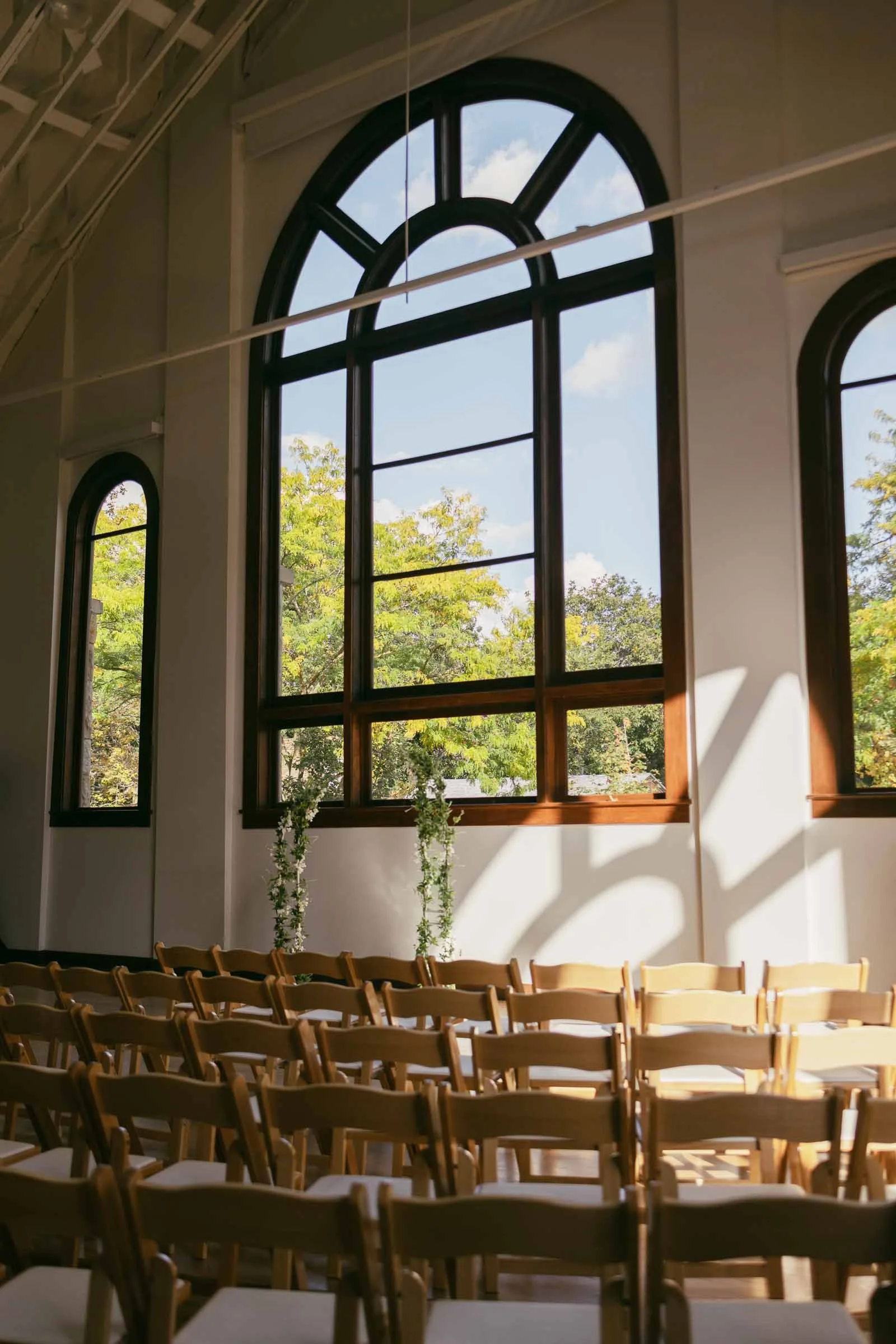  Rows of wooden chairs are arranged in front of large arched windows that let in sunlight, casting shadows on the floor. Outside, green trees are visible, creating a bright and peaceful indoor setting. 
