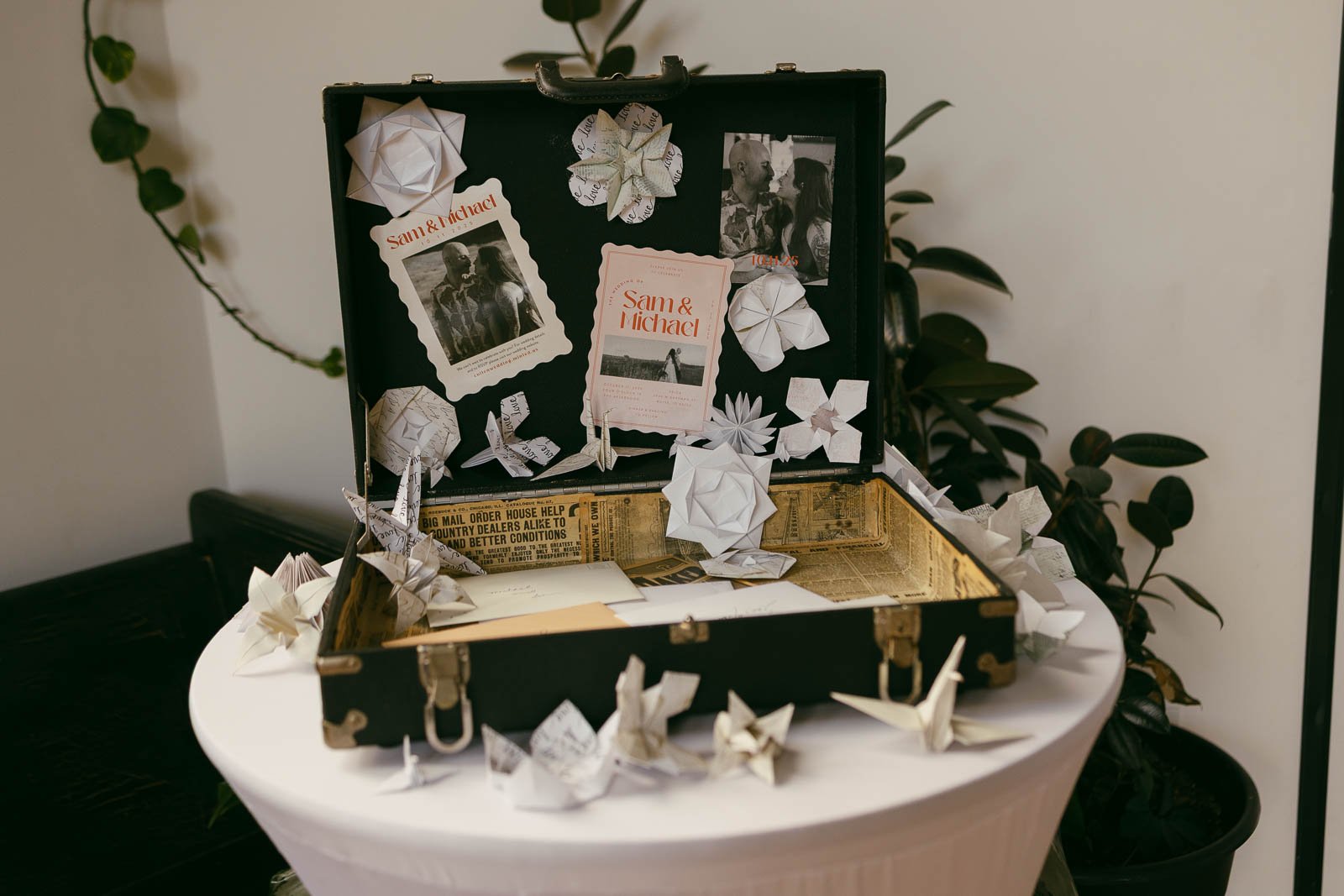  An open vintage suitcase decorated with origami flowers and paper cranes, photos, and cards that say "Sam &amp; Michael." The suitcase sits on a round table with plants in the background. 