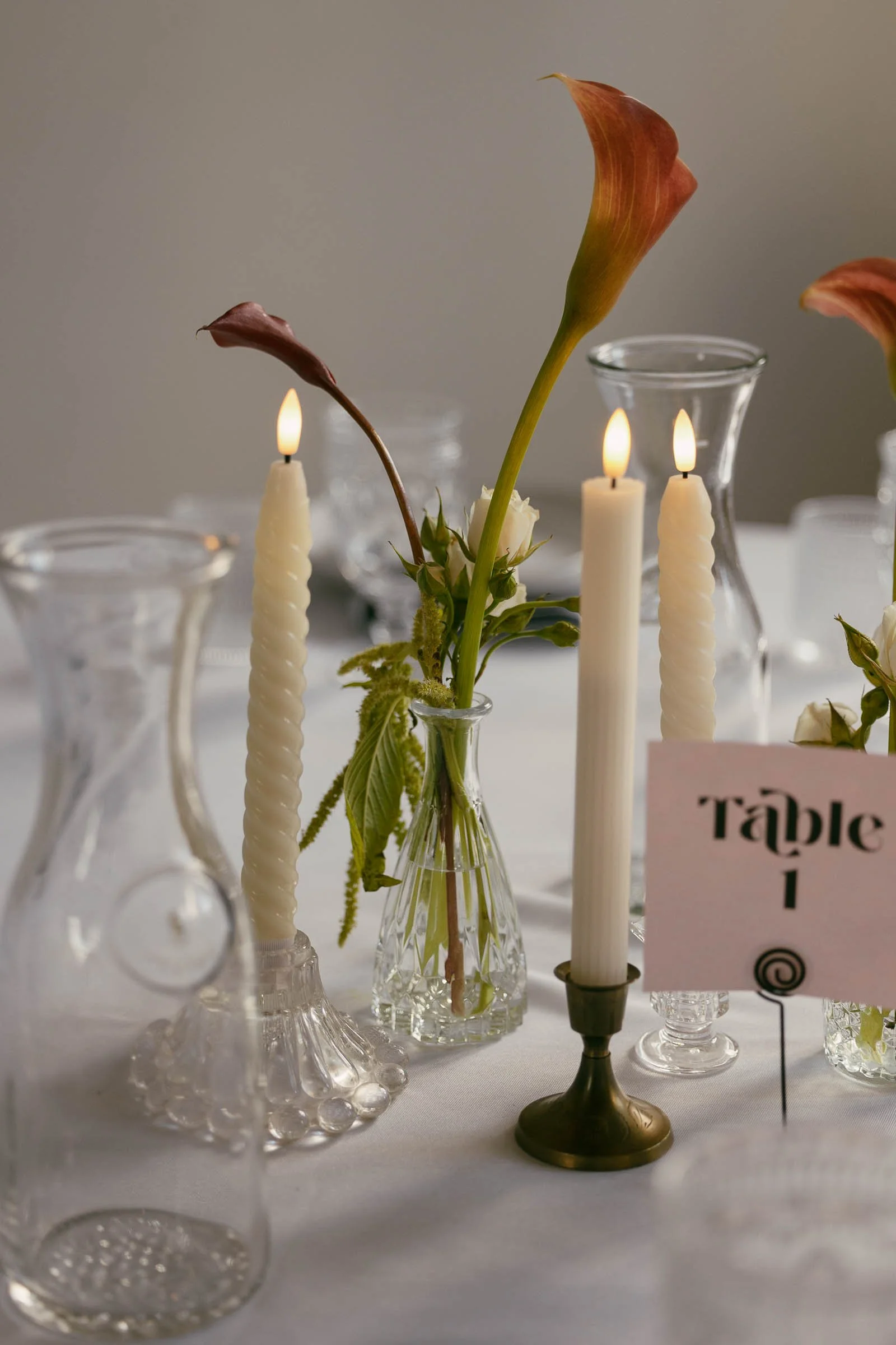  A close-up of a table centerpiece with two lit spiral candles, glass vases holding calla lilies and greenery, a "Table 1" card, and clear glass pitchers on a white tablecloth. 