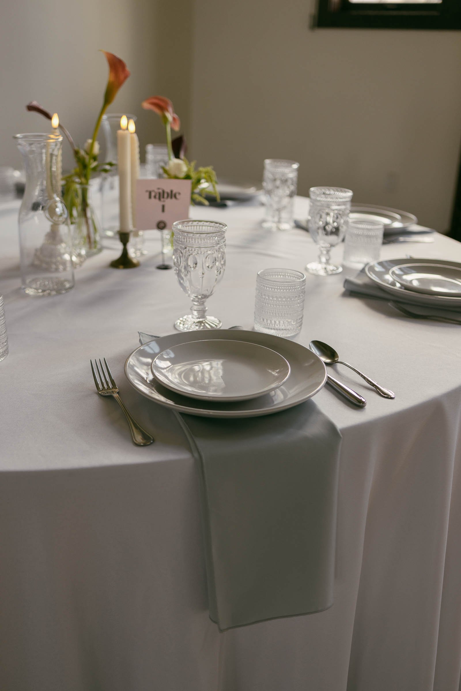  A round table set for a formal meal with white plates, silver cutlery, clear glasses, a pale gray napkin, candles, and simple flower arrangements on a white tablecloth. 