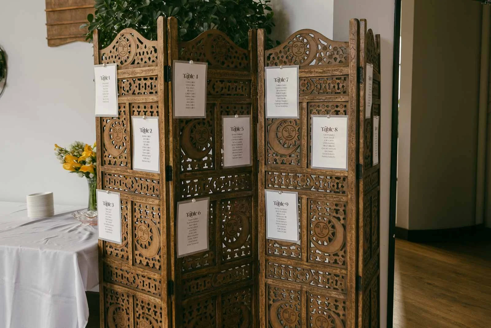  An ornate wooden folding screen displays white paper cards with table seating arrangements. The screen stands beside a table with a white tablecloth, plates, and a bouquet of yellow flowers. 