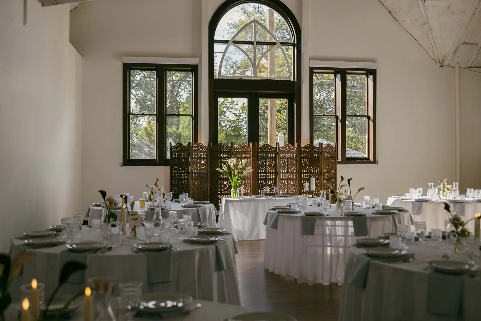  A bright, elegant room set for a formal event with round tables covered in white cloths, glassware, and floral centerpieces. Large arched windows and a decorative wooden screen are in the background. 