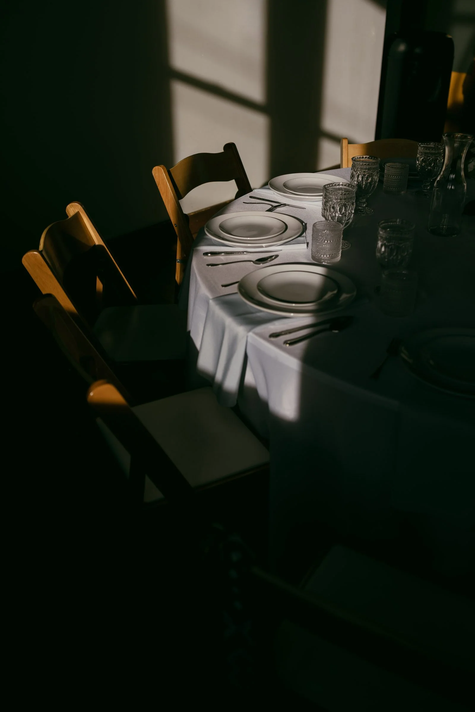  A round table set with white plates, glassware, and cutlery sits in partial sunlight, surrounded by wooden chairs. The light and shadows create a dramatic effect across the white tablecloth. 