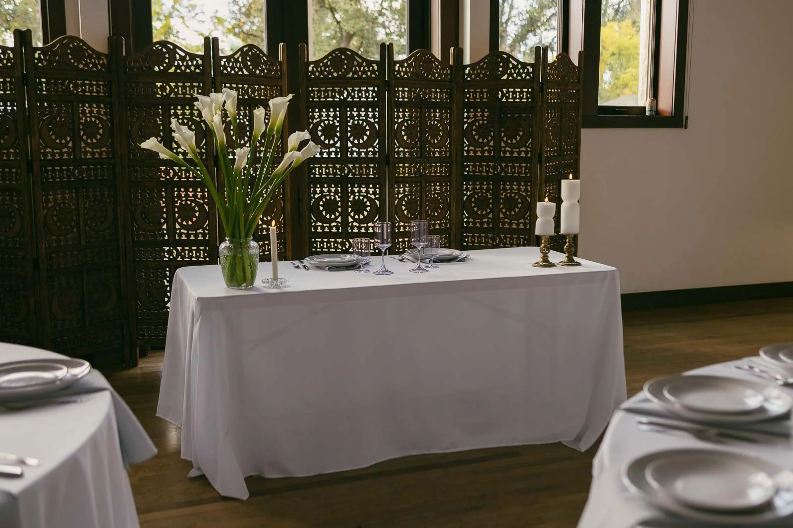  A white table with a white tablecloth is set for two, featuring calla lilies in a vase, candles, and glassware. Ornate folding screens and large windows are in the background, with more tables partially visible in the foreground. 