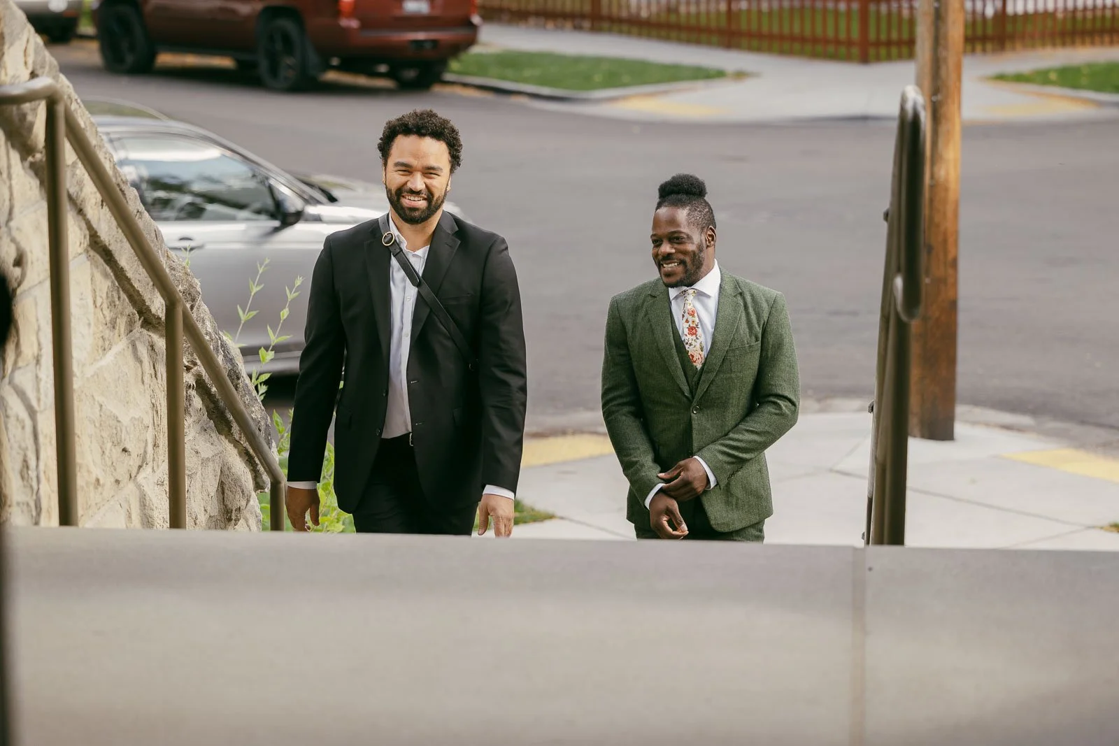  Two men dressed in suits are smiling and walking up outdoor steps on a city sidewalk. One wears a black suit, the other a green suit with a floral tie. Cars and a street are visible in the background. 