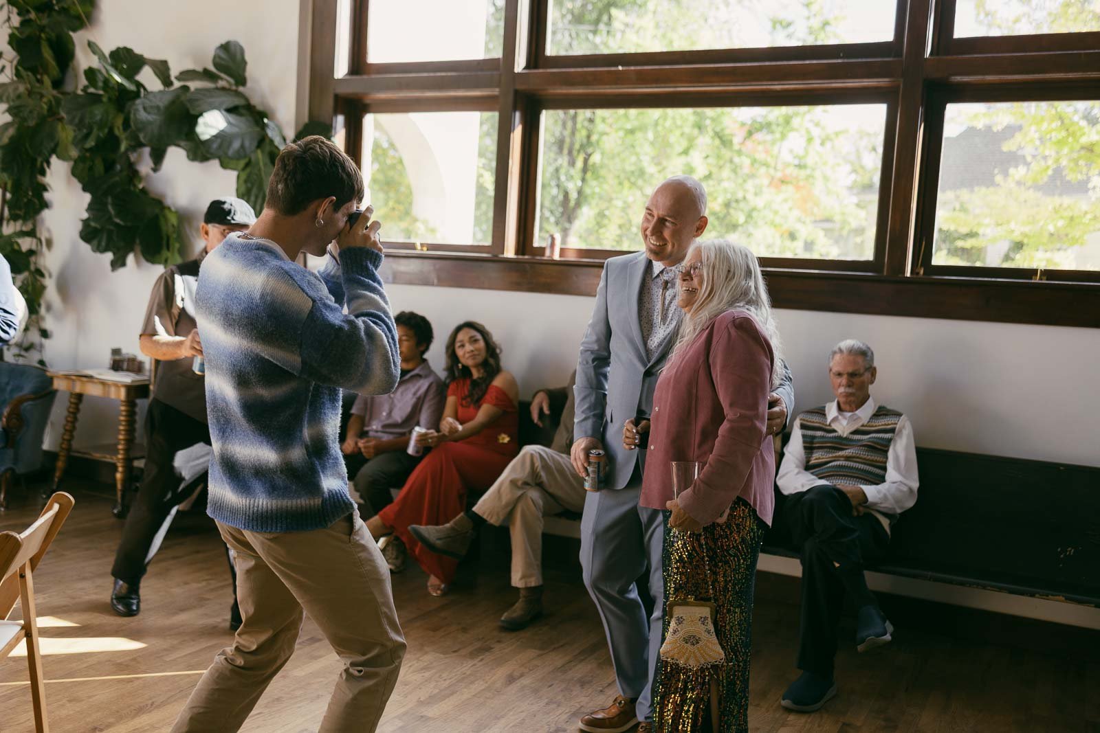  A man in a blue sweater takes a photo of a smiling couple posing together indoors, while several people sit and watch in a sunlit room with large windows and plants. 