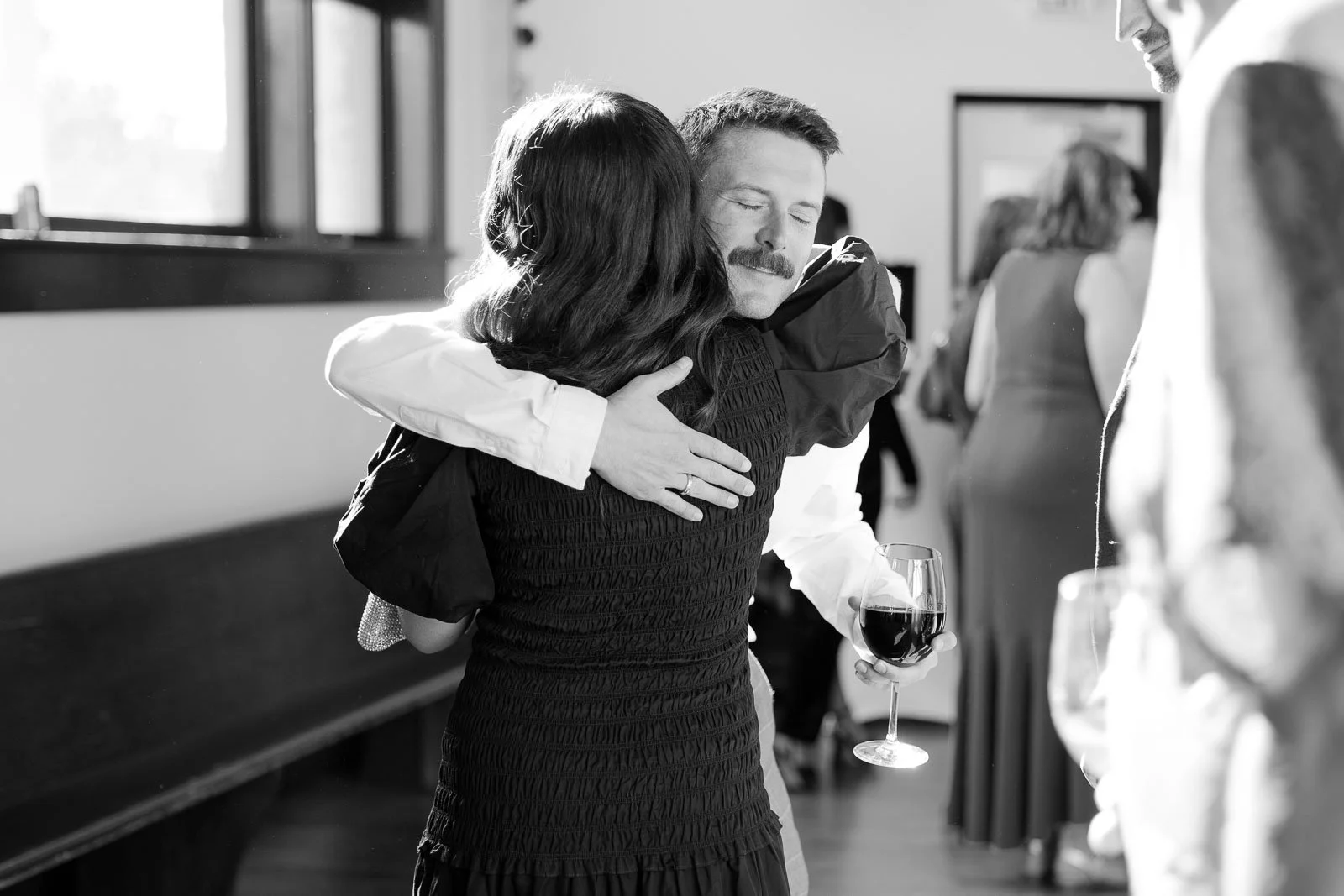  A man with a mustache hugs a woman, eyes closed and smiling, while holding a glass of wine. The scene is indoors with people in the background, captured in black and white. 