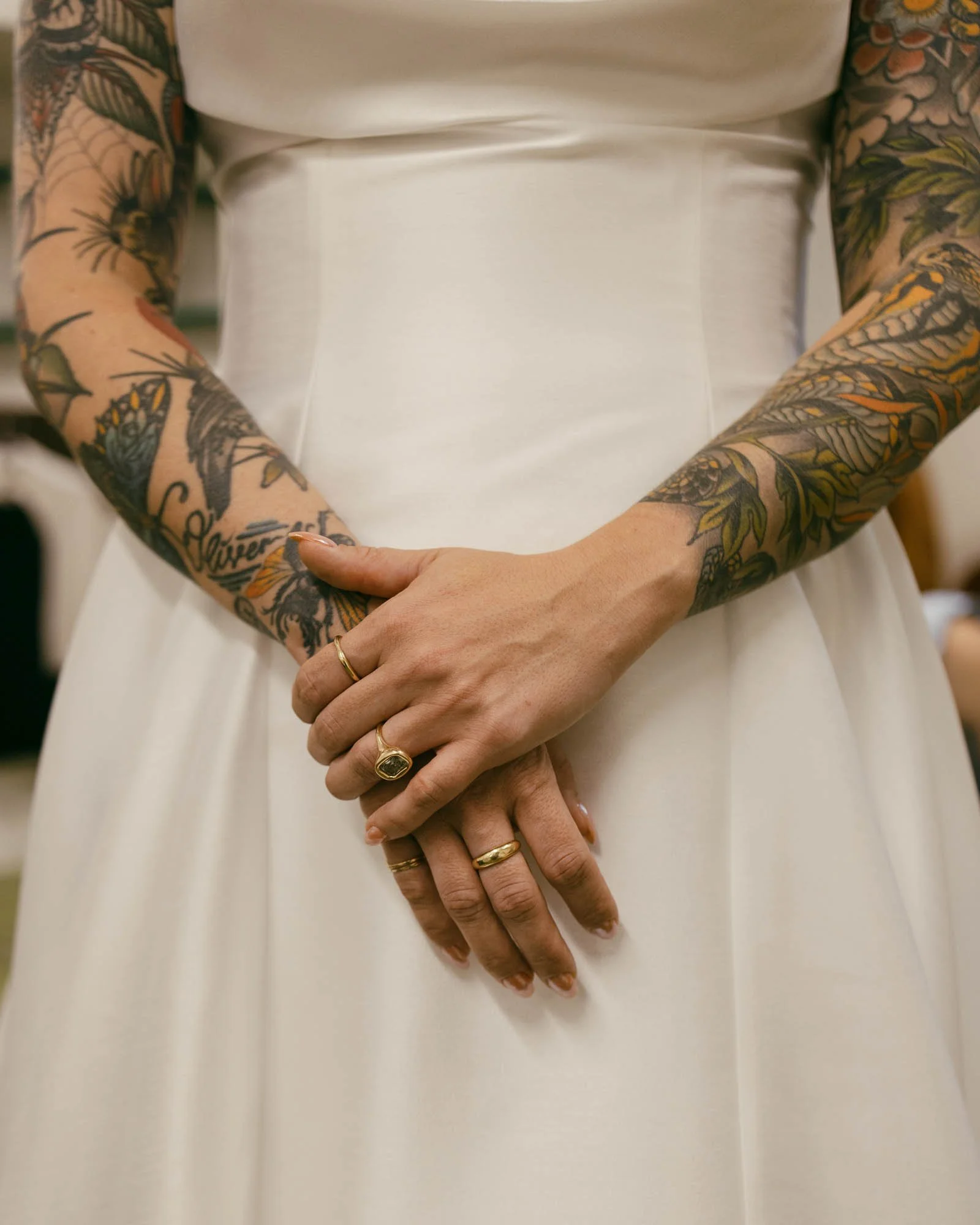  A woman in a white dress with tattooed arms stands with their hands clasped in front. She is wearing several gold rings and has intricate floral tattoos covering both arms. 