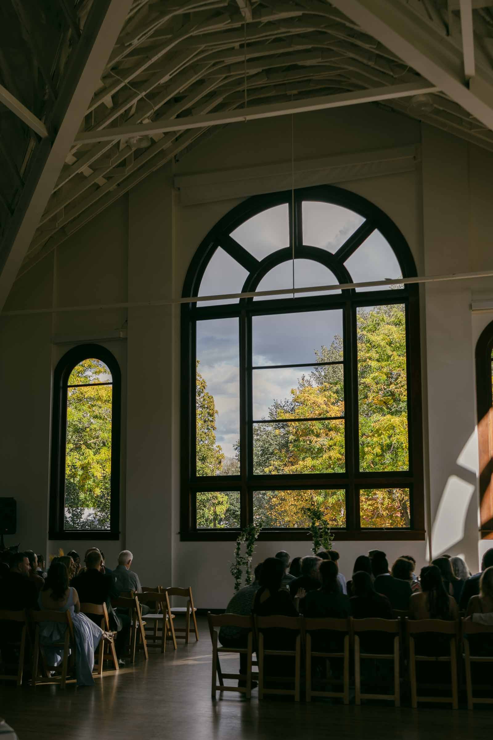  A group of people sit on wooden chairs indoors, facing large arched windows that reveal trees with green and yellow leaves outside. Sunlight streams through the windows, illuminating parts of the room. 