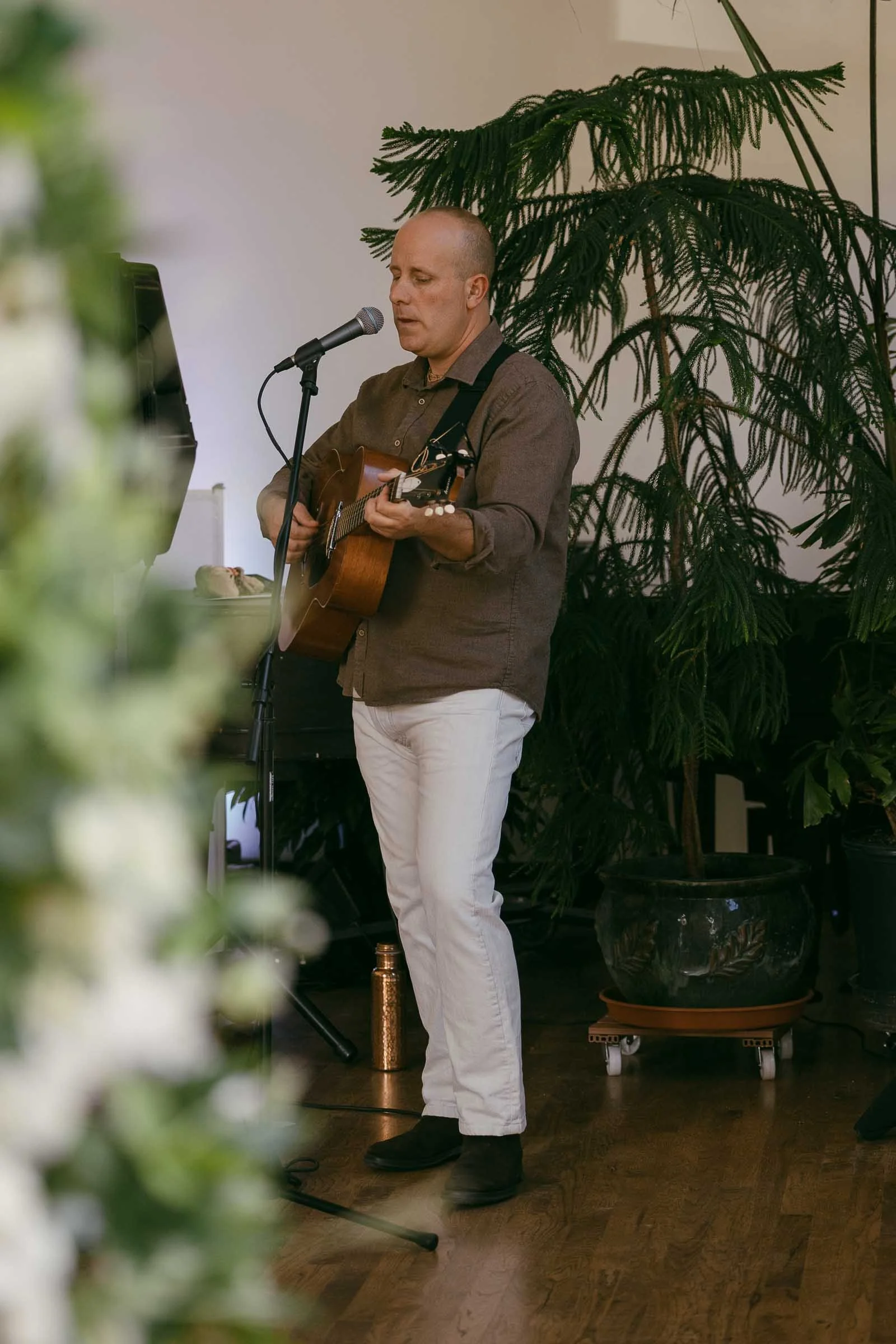  A man stands indoors playing an acoustic guitar and singing into a microphone, surrounded by green plants. He wears a brown shirt and white pants, and the floor is wooden. 