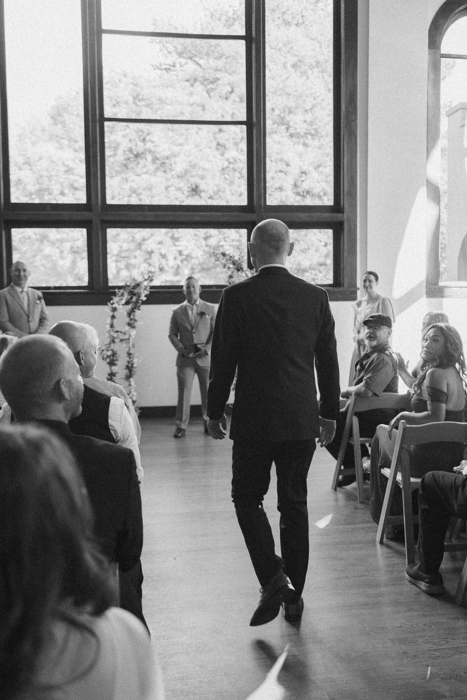  A man in a suit walks down the aisle of a sunlit room with large windows, while seated guests look on. The scene appears to be part of a formal event, possibly a wedding ceremony. 