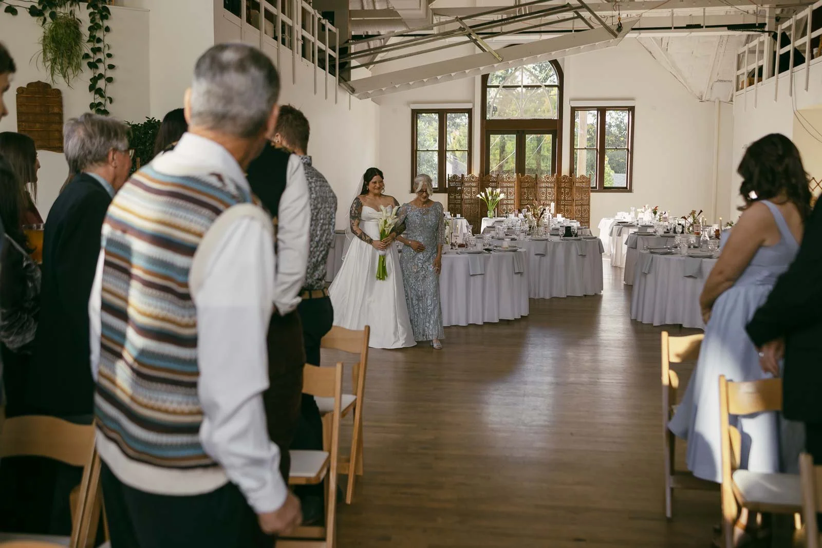  A bride in a white dress walks down the aisle with an older woman by her side, while guests in formal attire stand and watch in a decorated wedding venue with round tables and large windows. 