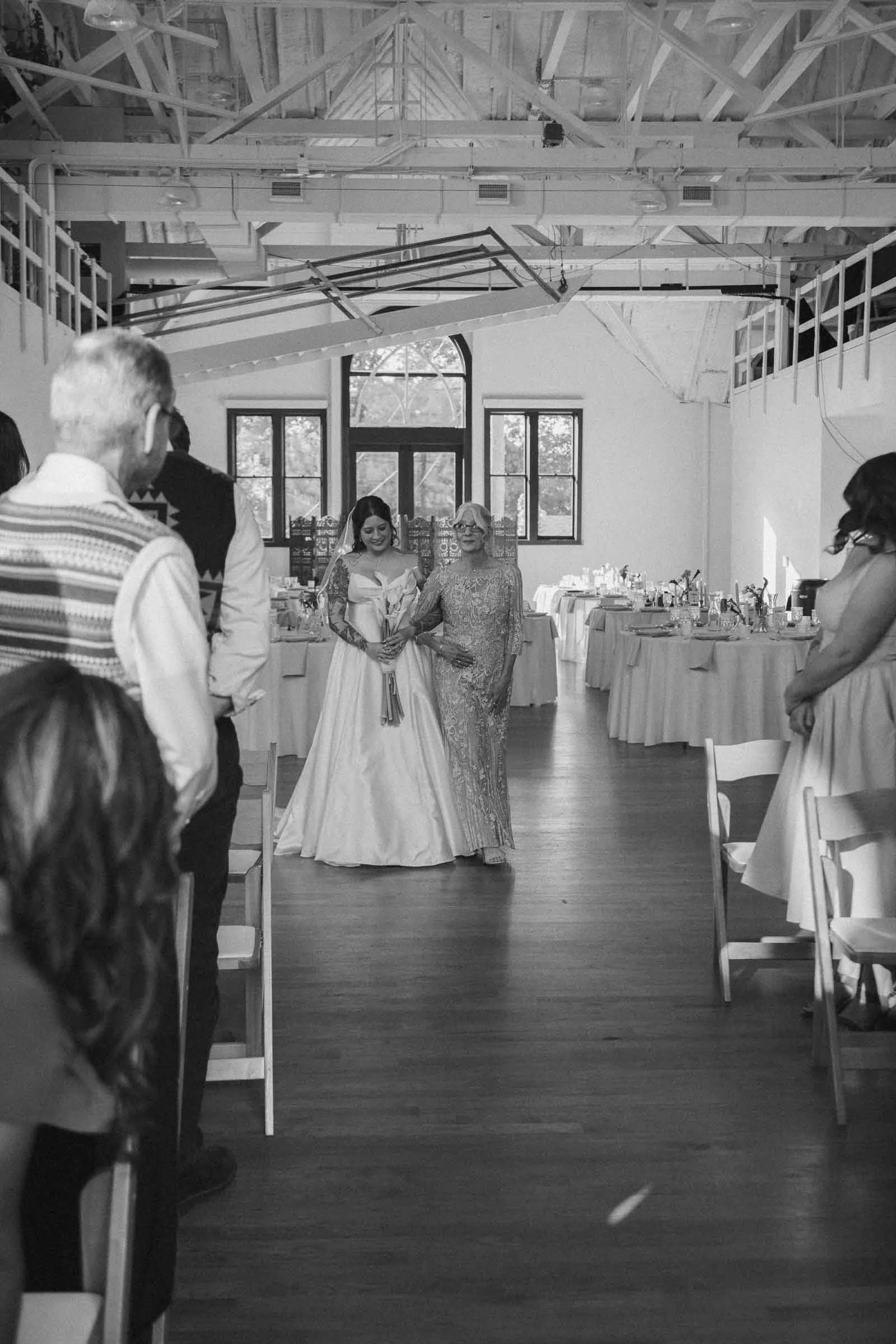  A bride in a gown holding a bouquet walks down the aisle with an older woman in a formal dress at a wedding ceremony, while seated guests watch in a decorated, light-filled venue. 