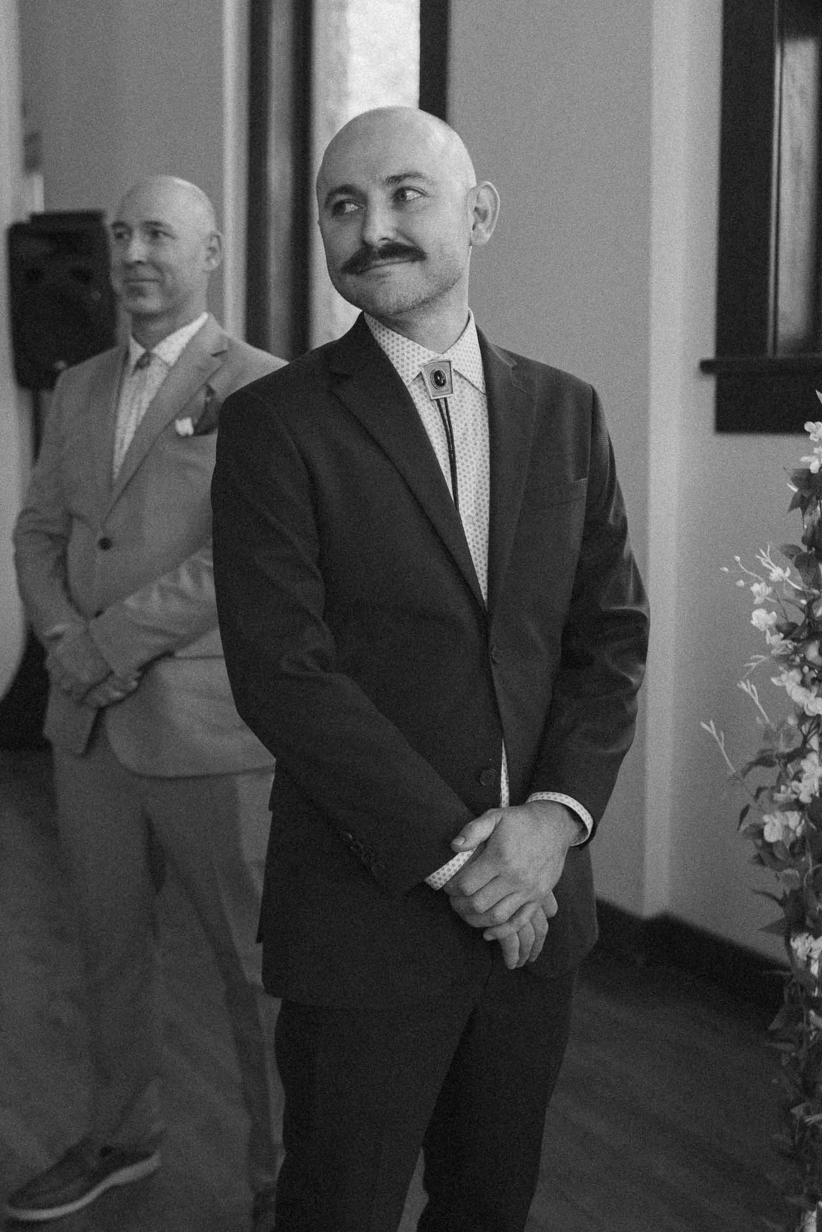  A man in a dark suit with a mustache stands smiling, hands clasped, indoors next to a floral arrangement. He has just seen his bride walk down the aisle 
