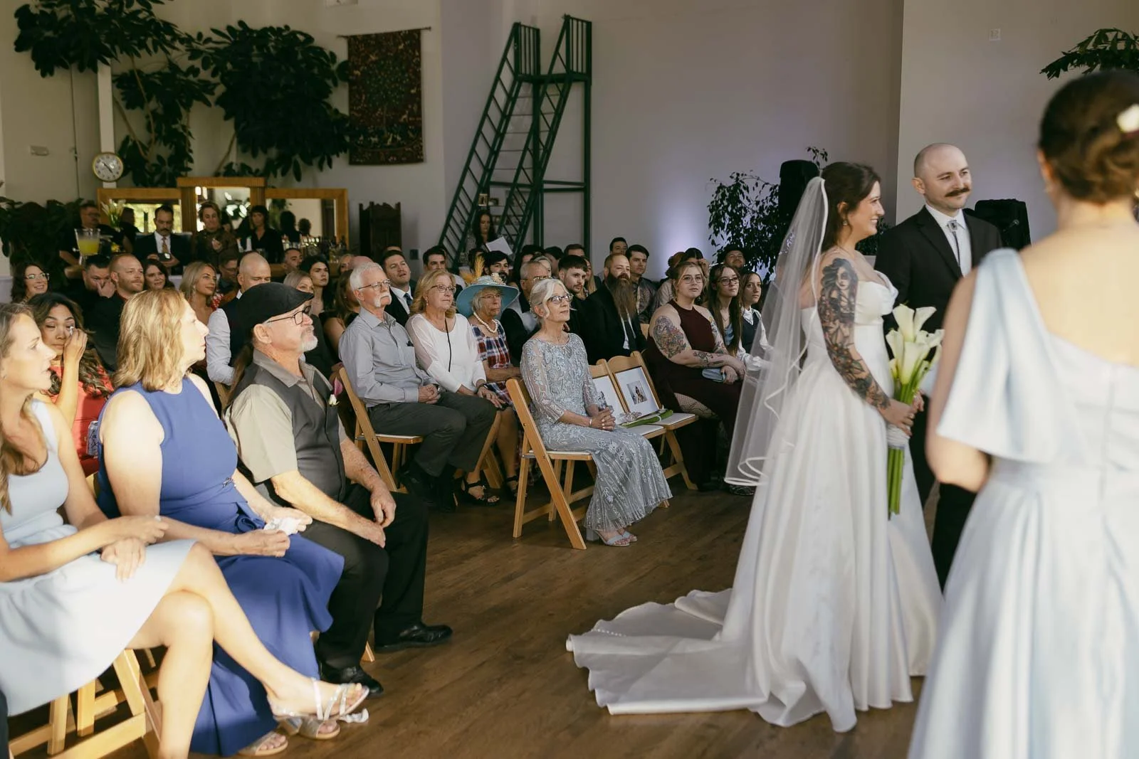  A bride and groom stand together at the front of a room filled with seated wedding guests. The guests are smiling and watching the ceremony. The venue is bright with natural light and decorated with green plants. 