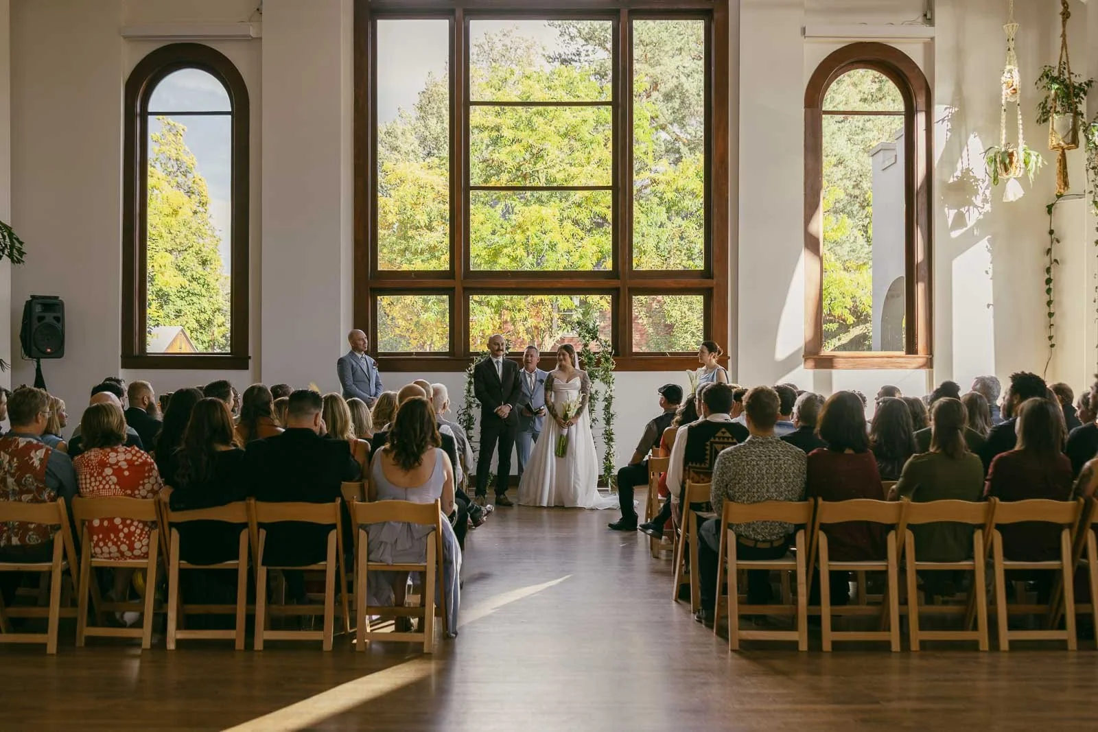  A wedding ceremony indoors with the bride and groom standing at the front, surrounded by family and guests seated in rows, large windows behind them showcasing green trees outside. 