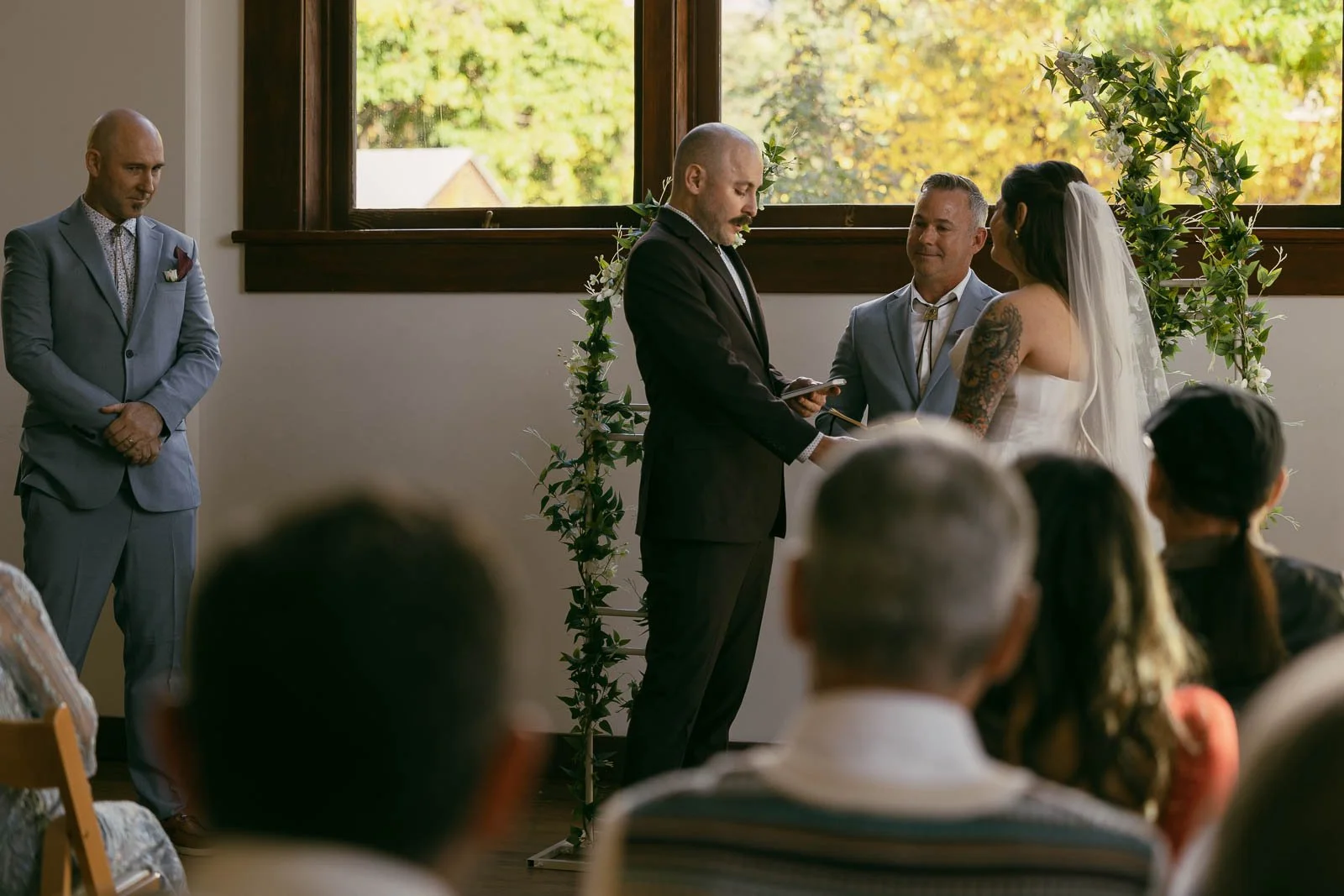  A couple stands holding hands at the altar during a wedding ceremony, with an officiant speaking and a man in a suit standing to the side. Guests are seated in the foreground, and large windows show trees outside. 