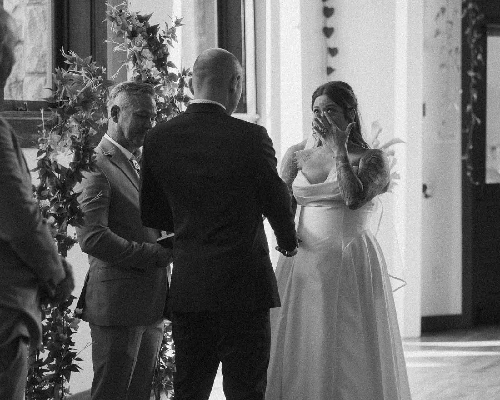 A bride in a white dress wipes away tears while holding hands with the groom during a wedding ceremony, with an officiant and floral decorations nearby. The scene is in black and white. 