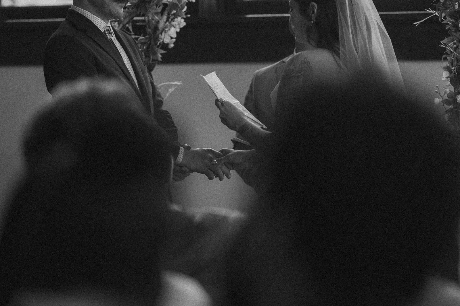  A black and white photo of a wedding ceremony, showing a couple holding hands as one reads vows from a piece of paper. The image is partially obscured by the blurred silhouettes of guests in the foreground. 