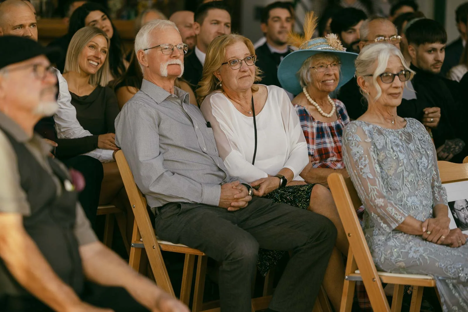  A group of adults, some smiling, sit closely together on wooden chairs at an indoor event. One woman wears a bright blue hat with feathers, while others are dressed in casual or semi-formal attire. 