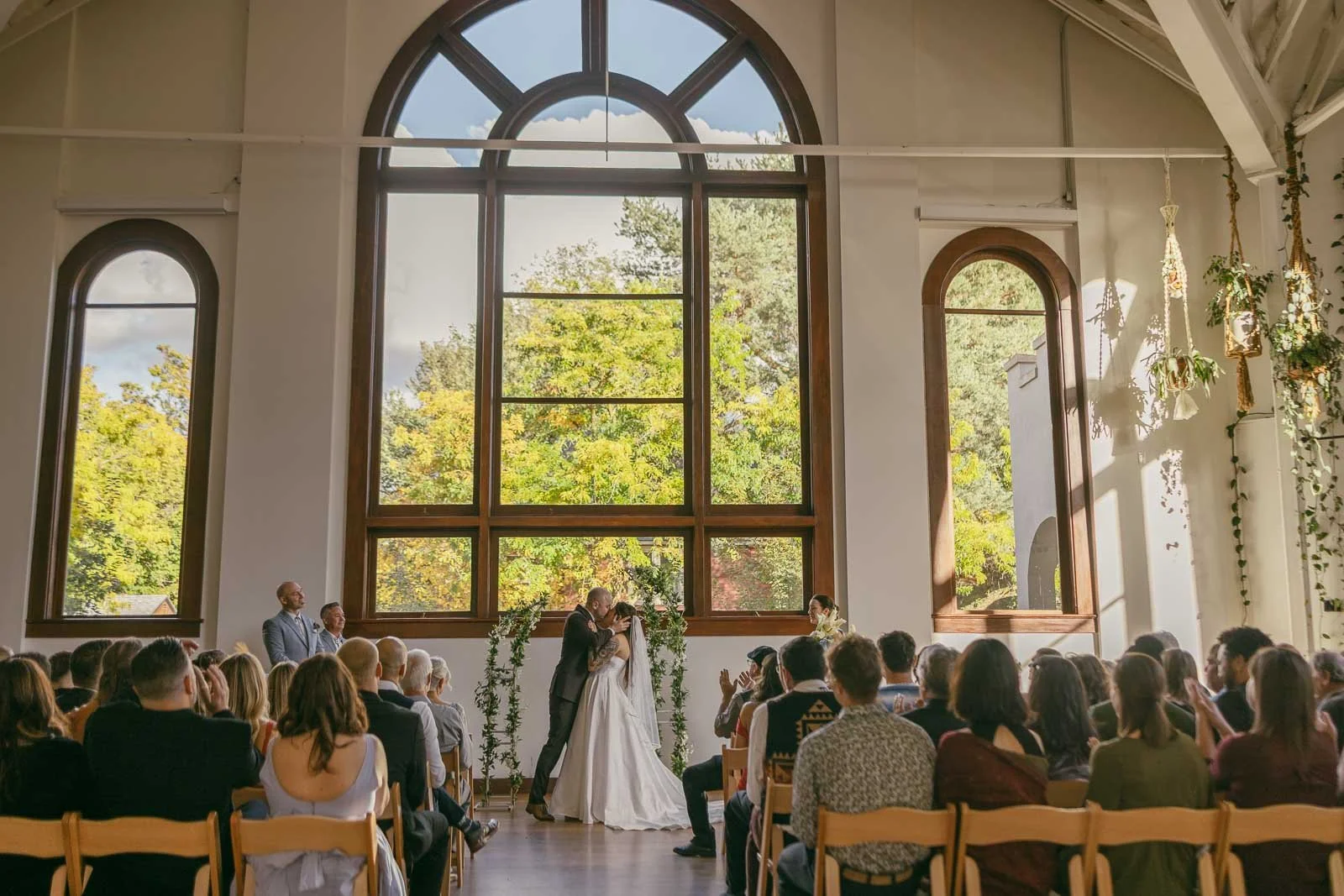  A couple shares a kiss at the altar during a wedding ceremony in a bright room with tall arched windows, surrounded by seated guests and lush greenery visible outside. 
