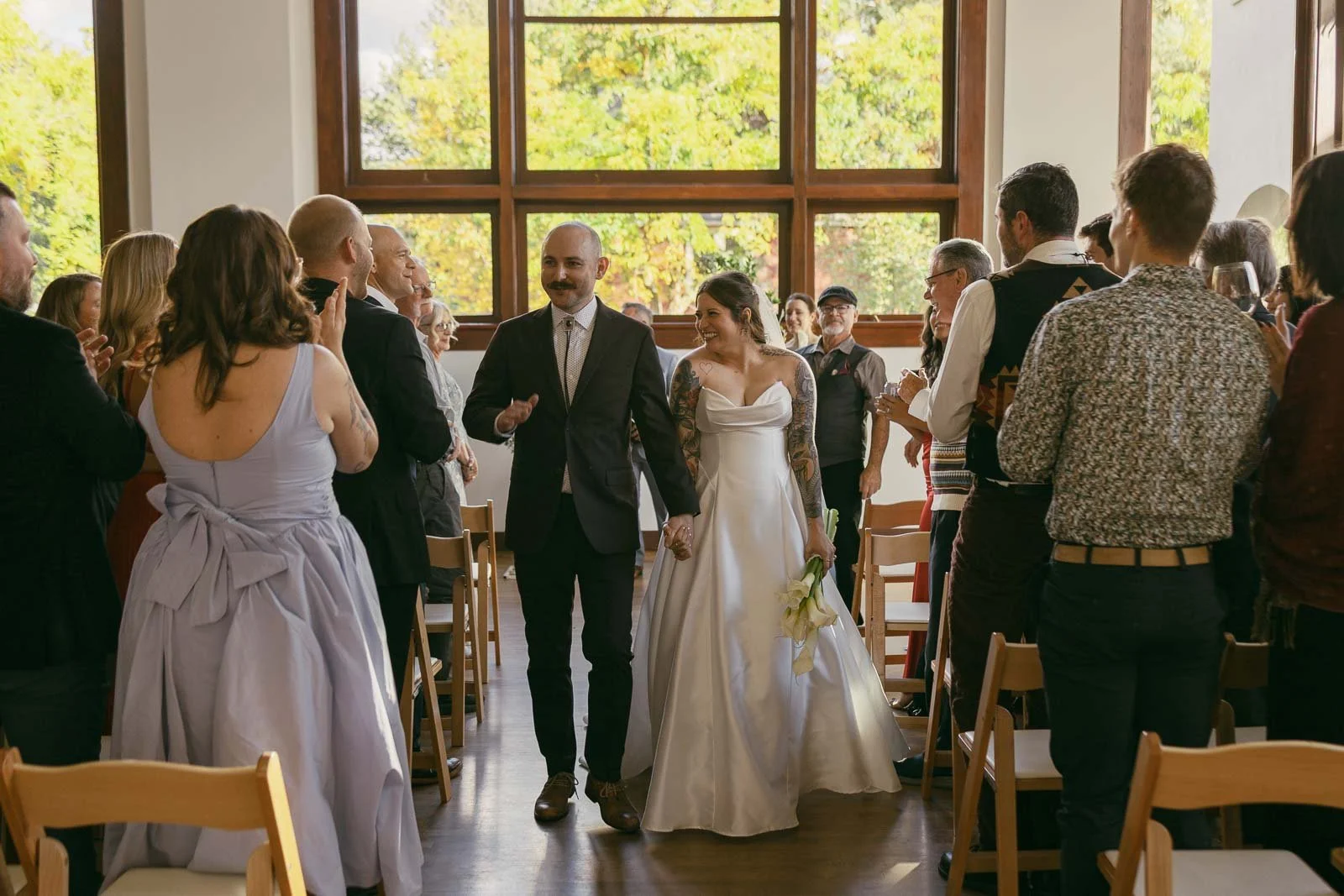  A newly married couple walks down the aisle, hand in hand, smiling as wedding guests stand and applaud. Sunlight streams through large windows in the background, illuminating the joyful scene. 