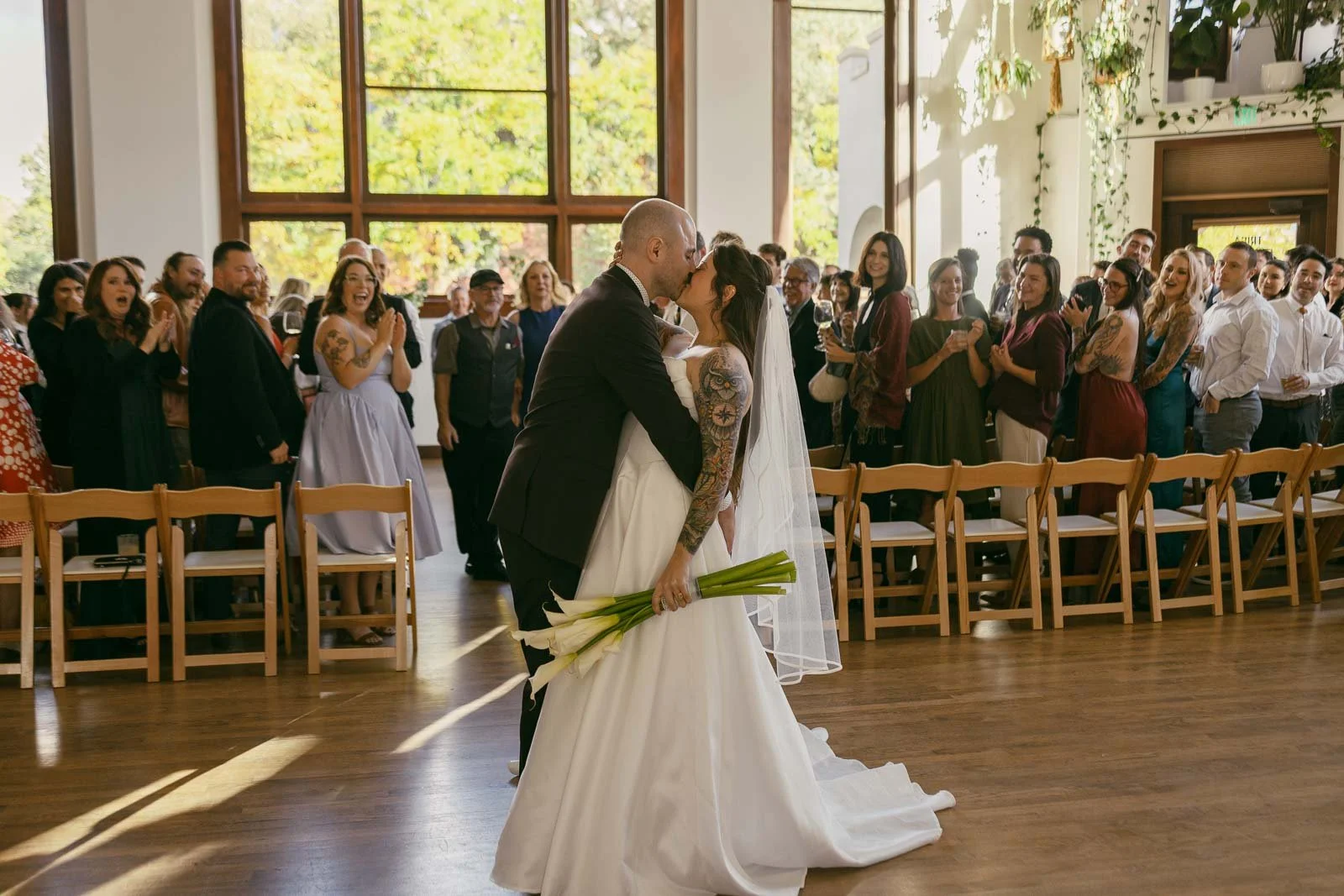 A just-married couple shares a kiss at the end of the ceremony aisle inside a bright, sun-filled room as their guests cheer and applaud around them. The bride is holding a bouquet of white calla lilies