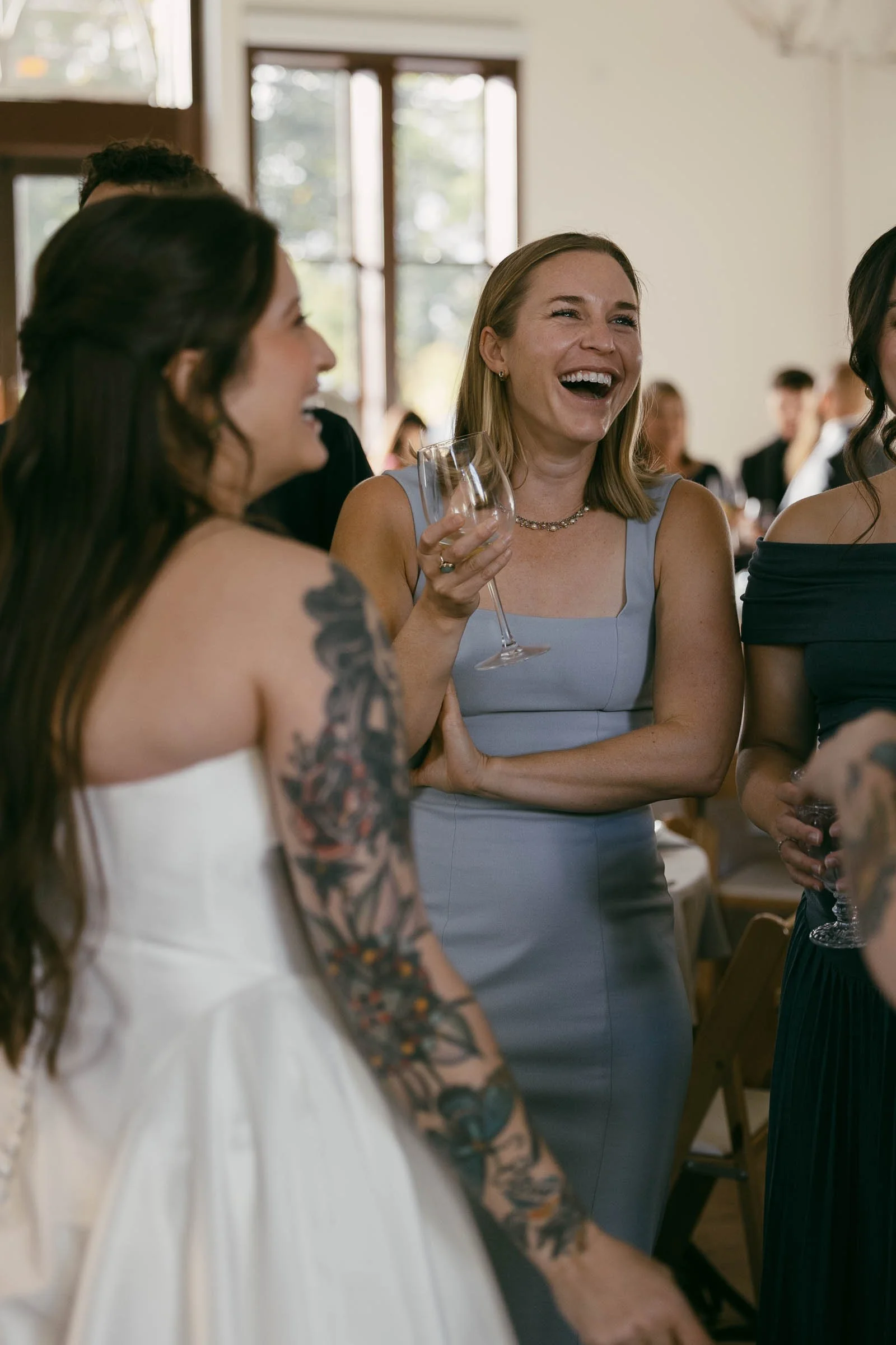  A group of women, including a bride in a white dress, stand together indoors, laughing and smiling. One woman in a light blue dress holds a wine glass. The setting appears to be a wedding reception. 