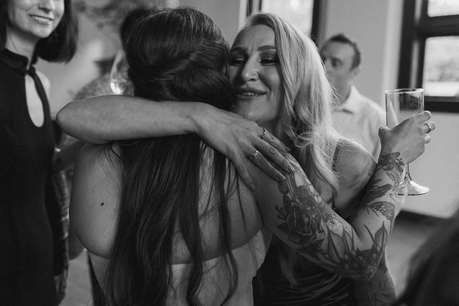  A woman with long hair and tattoos smiles while hugging another woman at a gathering. She holds a champagne glass in one hand, and other people are visible in the background. The image is in black and white. 