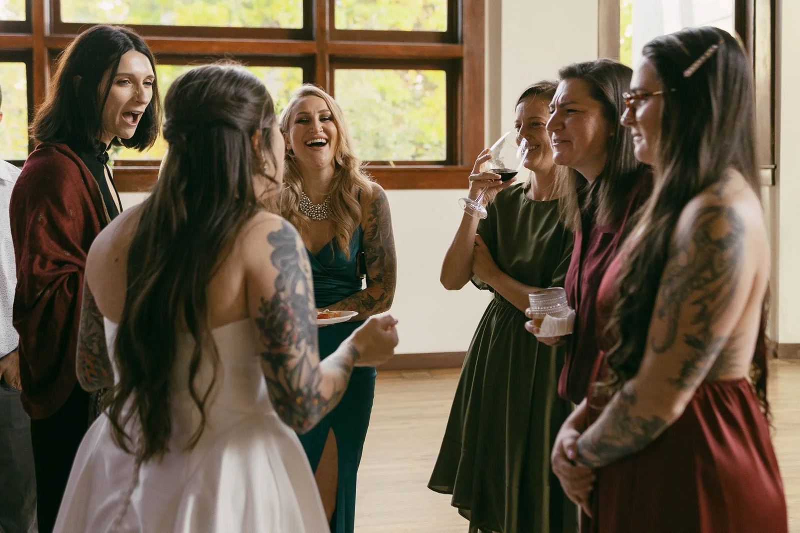  A bride in a white dress stands with a group of women, all smiling and laughing together indoors. The women are dressed in colorful dresses and some have visible arm tattoos. Sunlight streams through large windows behind them. 