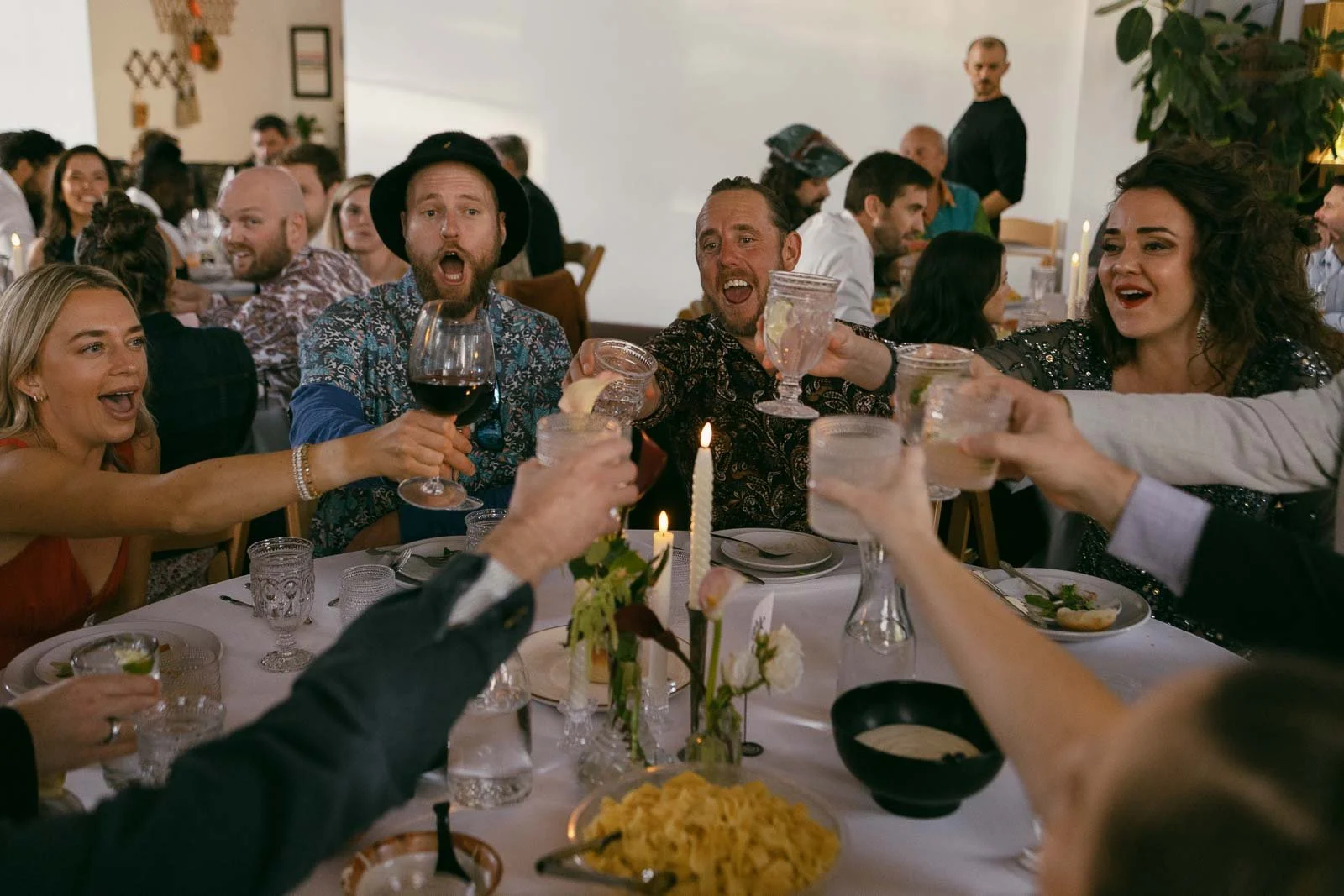  A group of people seated around a table raise their glasses in a celebratory toast during a festive gathering or party, smiling and enjoying the moment. The table is set with food, drinks, and candles. 