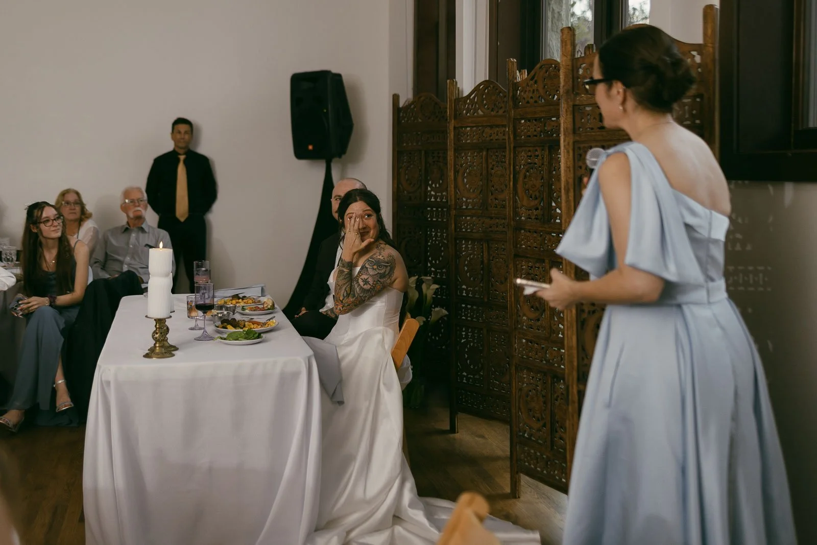  A bride in a white dress wipes away tears while seated at a table, listening to a woman in a light blue dress giving a speech. Guests watch attentively in a warmly lit reception room. 