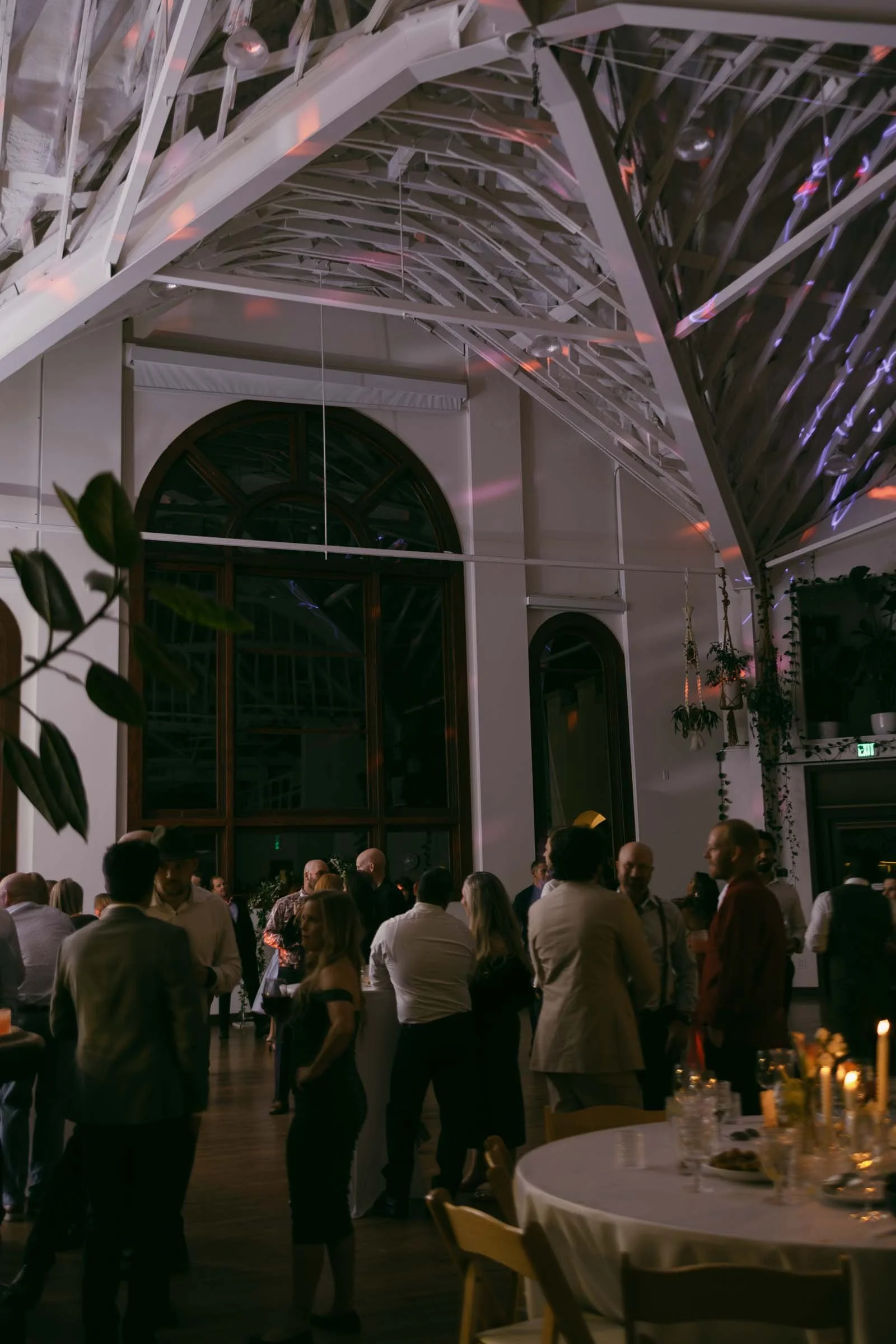  Guests mingle and chat in a warmly lit venue with high ceilings, large arched windows, hanging plants, and candles on tables, creating an inviting atmosphere for a social event or celebration. 