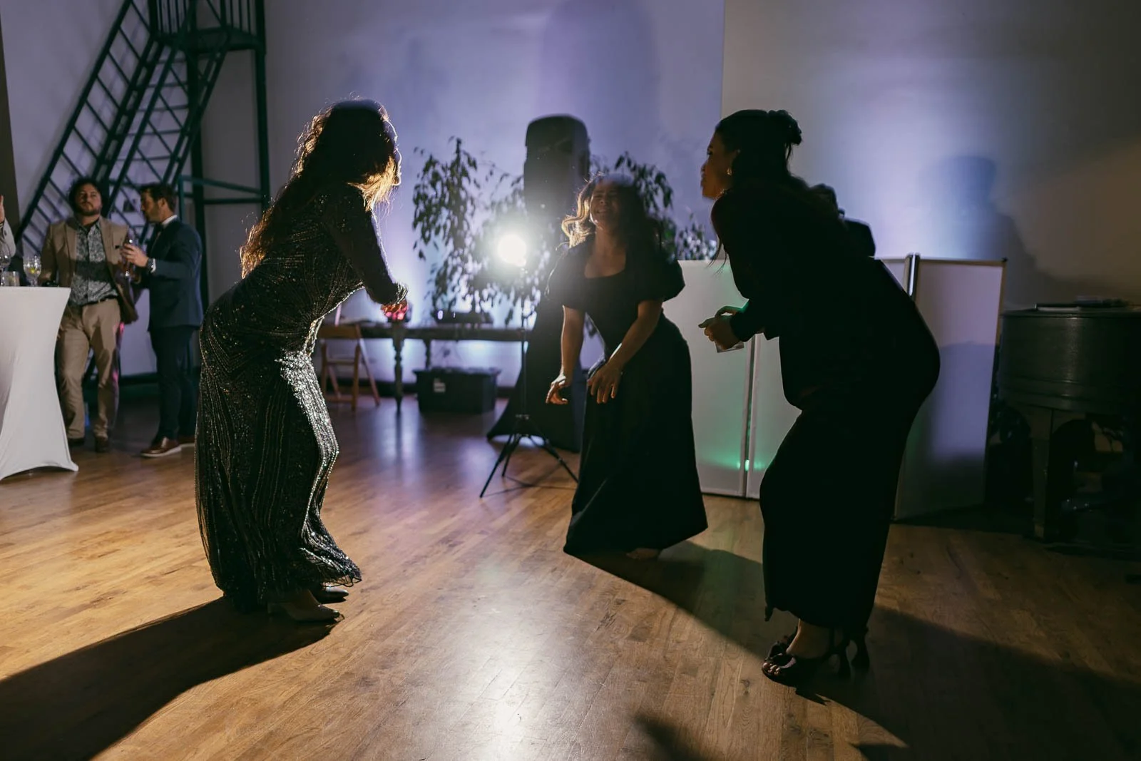  Three women in formal dresses dance energetically on a wooden floor at a party, with dramatic lighting and other guests and equipment in the background. 