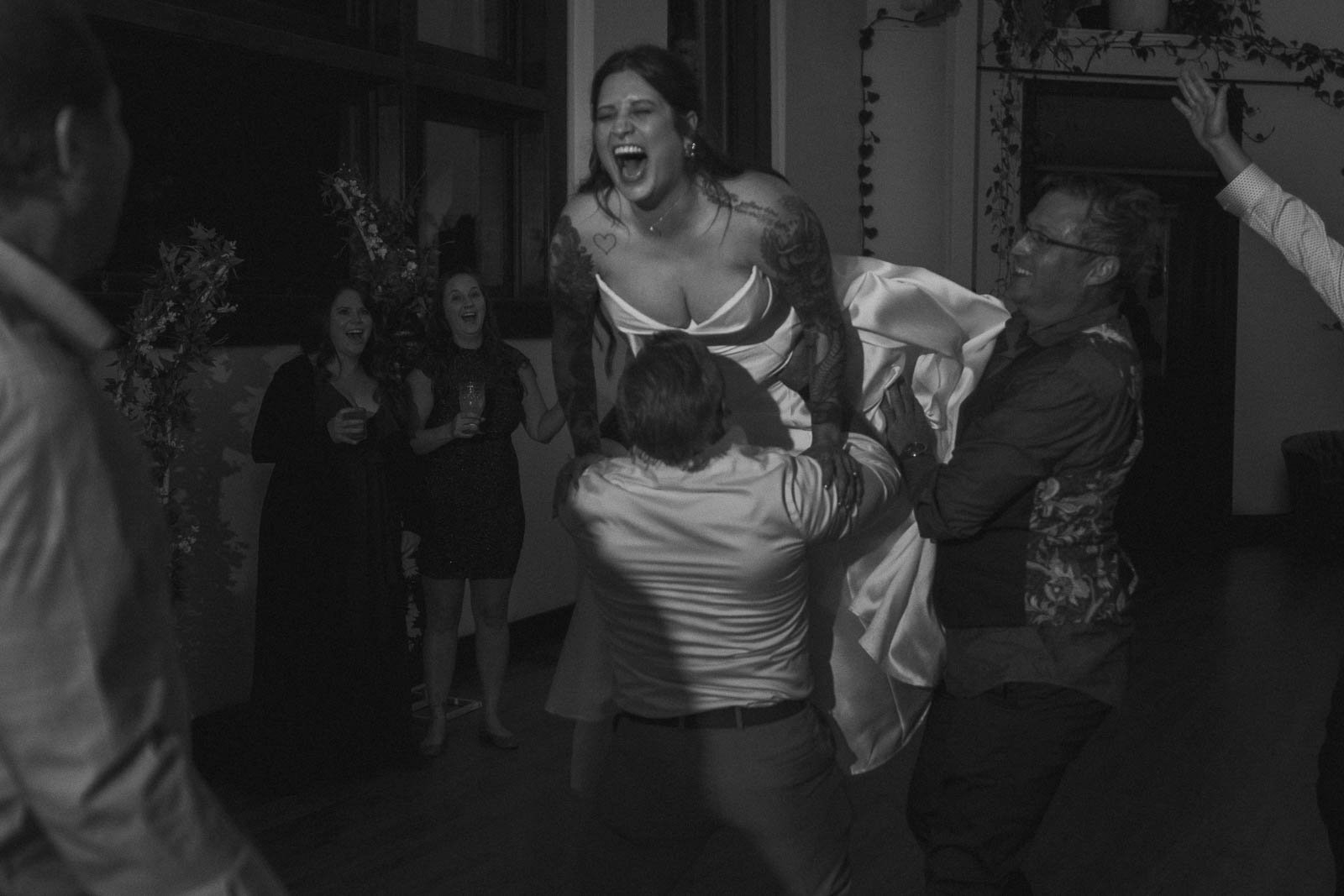  A joyful bride in a wedding dress is lifted in the air by two guests at a lively indoor celebration, with onlookers smiling and cheering in the background. The scene is captured in black and white. 
