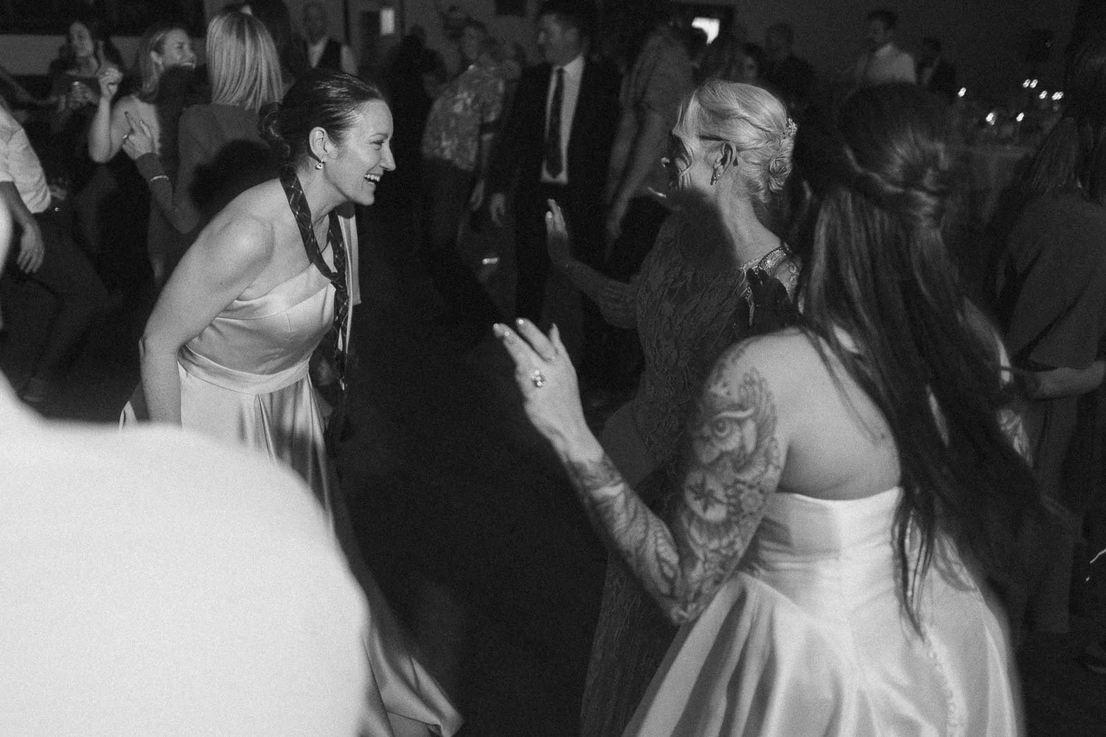  A group of women in dresses are dancing and smiling at a lively indoor event. One woman has visible tattoos on her arm. Other people are mingling and dancing in the background. The image is in black and white. 