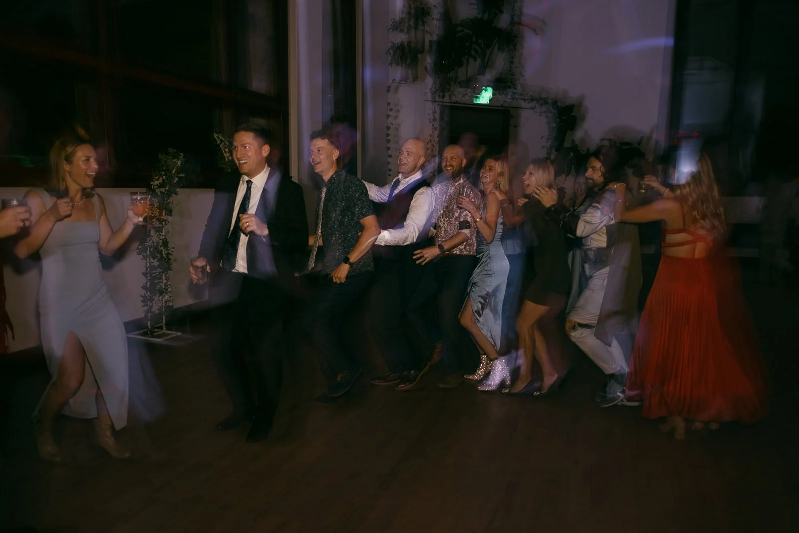  A group of people dressed in formal attire dance in a conga line at an indoor event, smiling and laughing, with blurred motion capturing their energetic movement. 