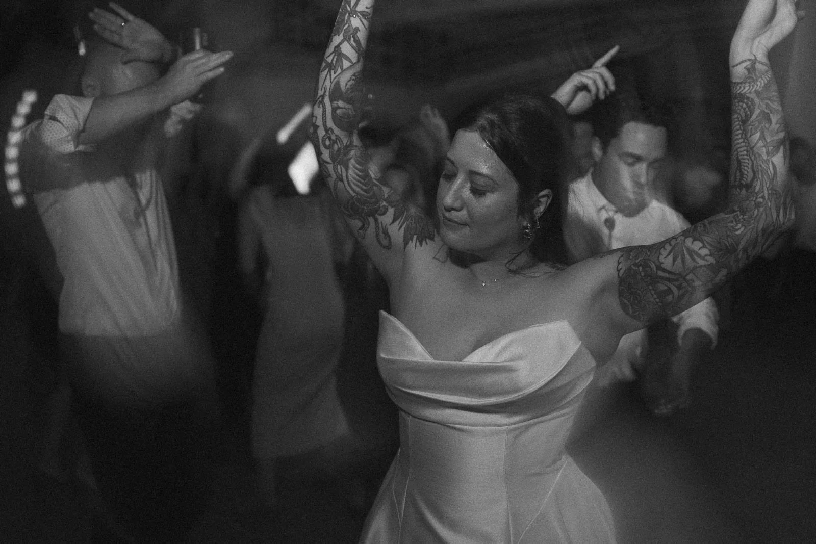  A woman in a strapless dress with tattooed arms dances with her eyes closed, surrounded by other people dancing at a dimly lit event or party. 