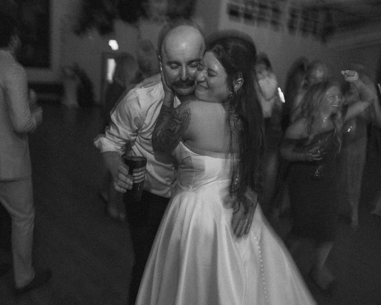  A couple embraces and smiles while dancing at a lively indoor event. The woman wears a strapless dress and the man holds a drink. Other guests dance and celebrate in the softly lit background. 