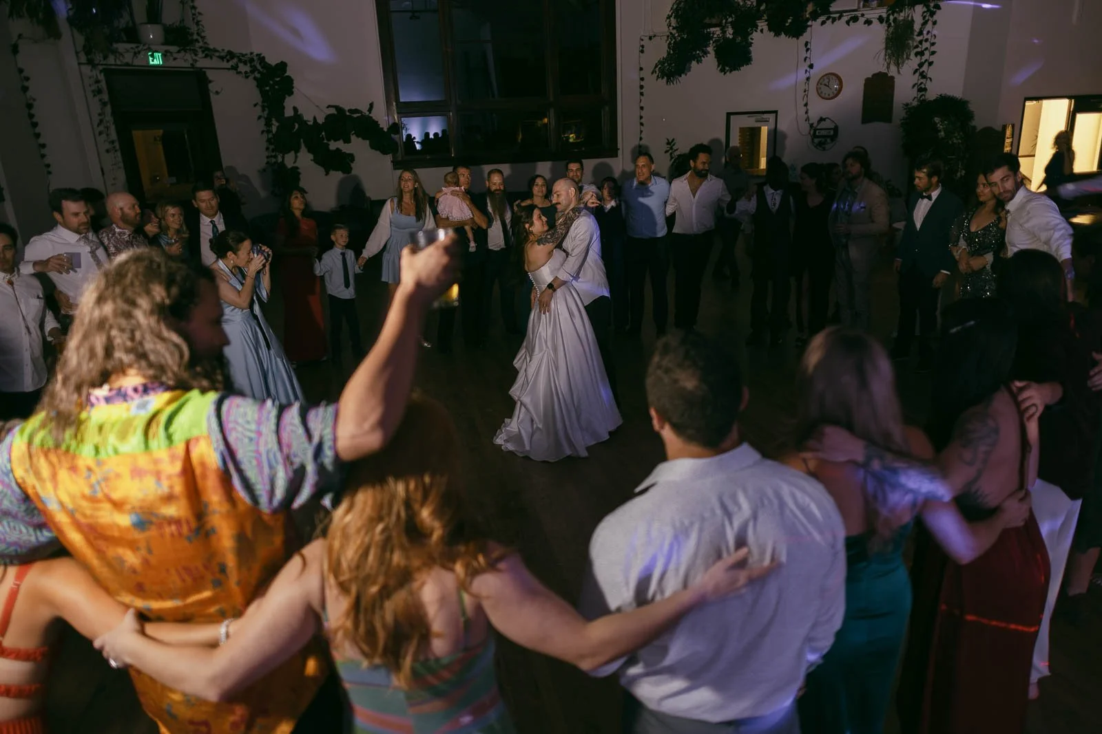  A bride and groom share their first dance in the center of a room, surrounded by guests standing in a circle with arms around each other, watching and celebrating the couple. The atmosphere is festive and joyful. 