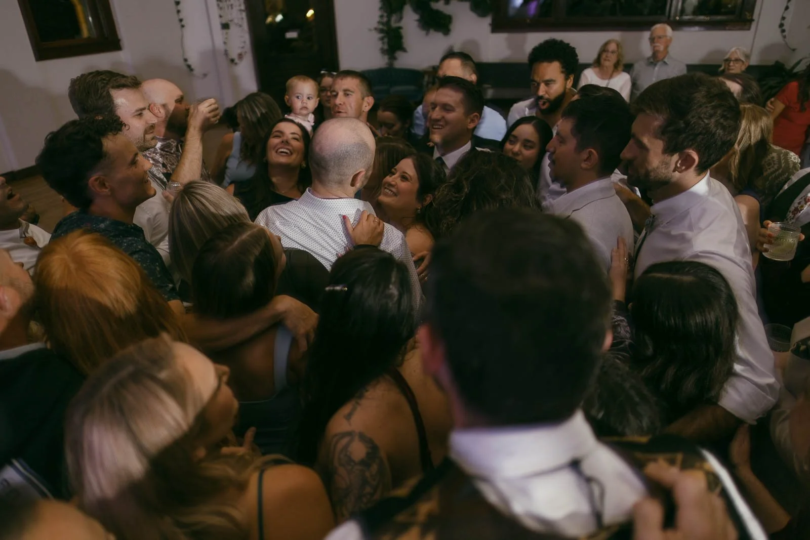  A large group of people gather closely together indoors, smiling and laughing, suggesting a joyful celebration or party. Some people have their arms around each other, creating a warm, festive atmosphere. 