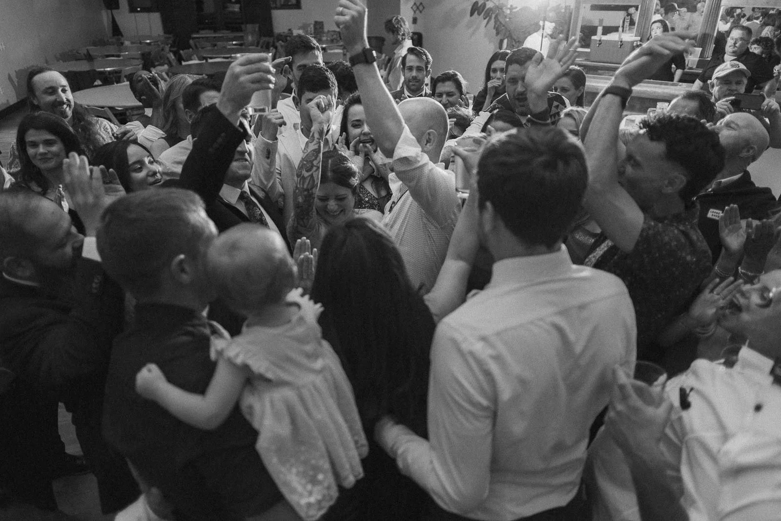 A black and white photograph shows a mixed group of men, women, and a child standing close together at an energetic indoor celebration, lifting their hands and raising glasses in a joyful toast.