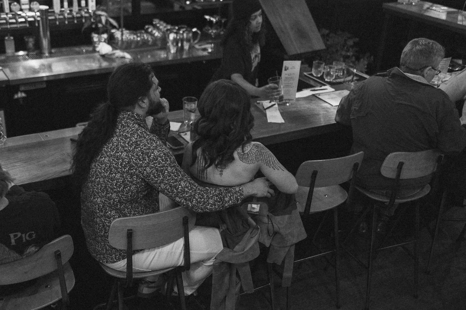  A man and woman sit closely together at a bar, with the man's arm around the woman's waist. Other patrons sit nearby on barstools, and a bartender works behind the counter. The scene is in black and white. 