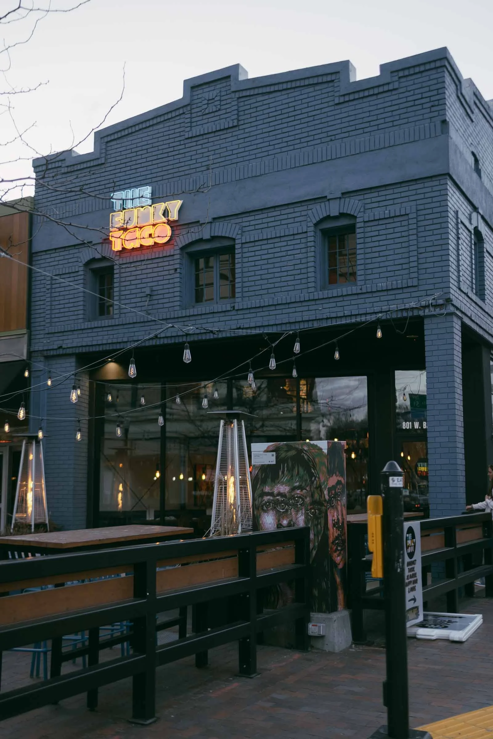  A blue brick restaurant called "The Funky Taco" with a neon sign above the entrance, outdoor seating, string lights, patio heaters, and a painted mural near the patio. 