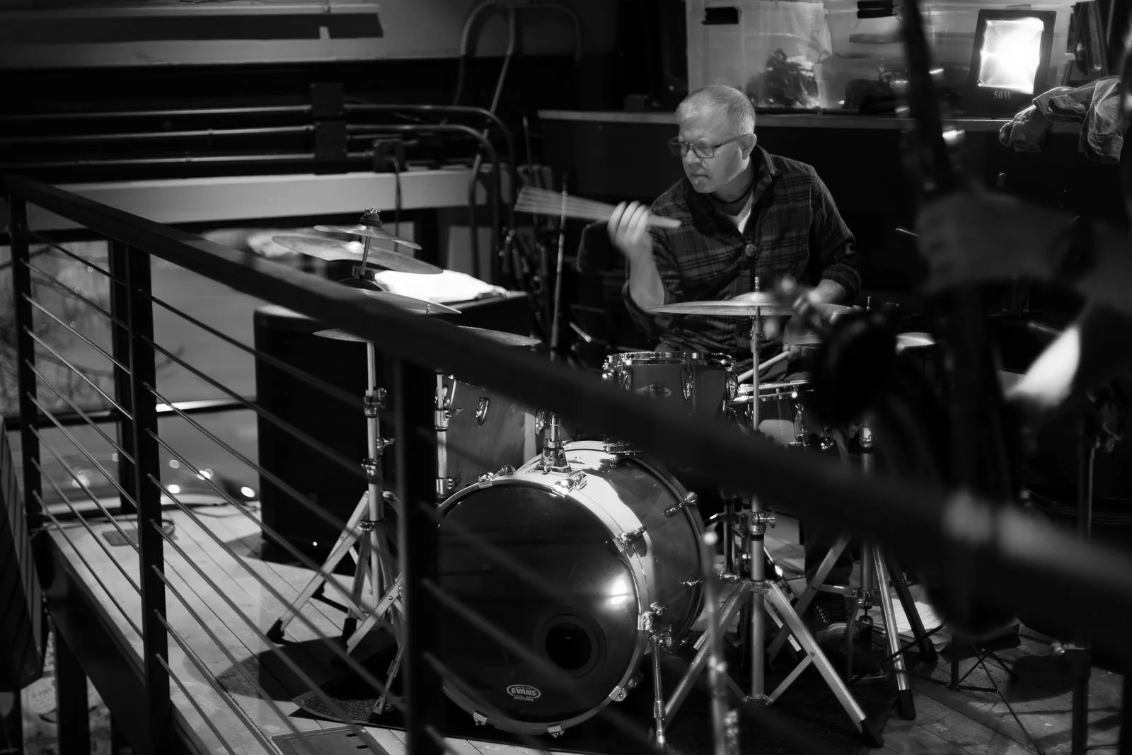  A man plays a drum set intensely in a dimly lit room, partially obscured by metal railings in the foreground. The scene is in black and white, capturing a focused moment during a live performance. 