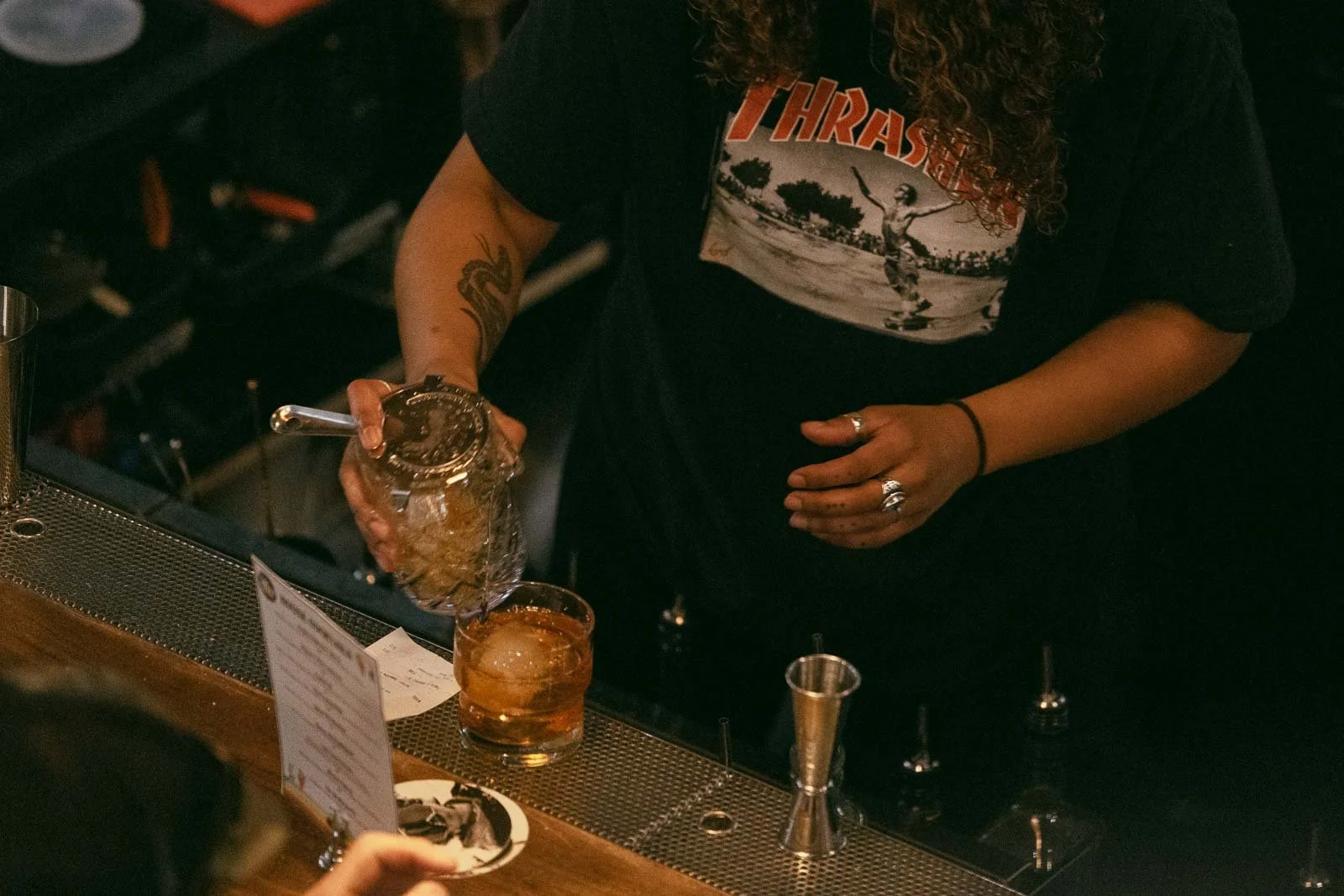  A bartender wearing a black "Thrasher" t-shirt pours ice from a jar into a glass on a bar counter, surrounded by bartending tools and a cocktail menu. 