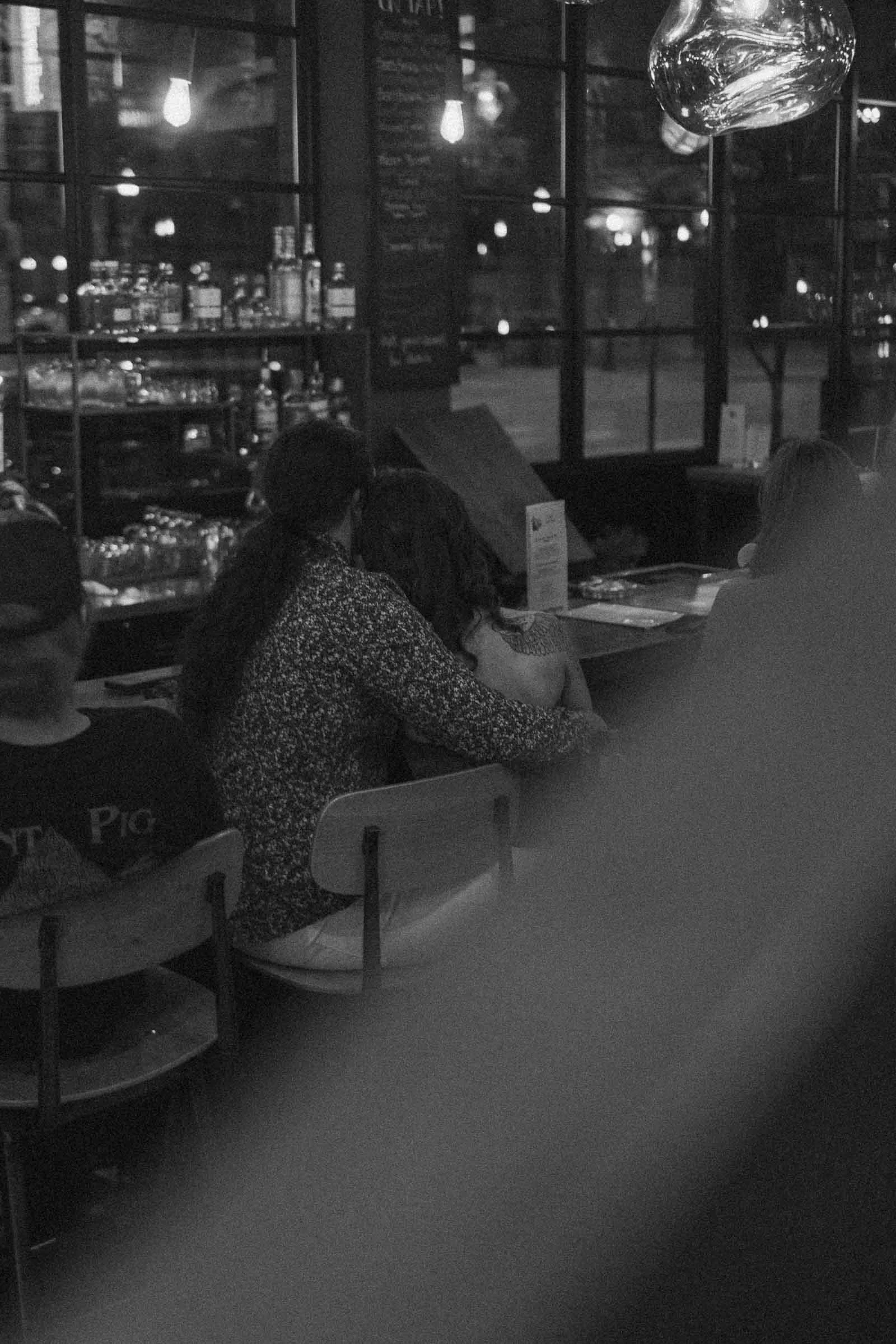 A black and white photo shows a couple sitting closely together at a bar, illuminated by dim lighting. Bottles and glasses line the shelves behind the bar, and other patrons sit nearby. It is Jazz Night at Funky Taco. 